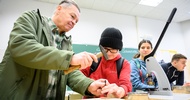 Eine Person und ein Kind stehen nebeneinander an einem Tisch. Die Person hilft dem Kind einen Nagel in ein flaches, rundes, kleines Holzstück zu schlagen