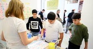 Zwei Personen stehen sich an einem Tisch gegenüber. Die rechte Person malt mit einer Malerrolle auf einem vor ihr liegenden Stück Papier verschiedene Muster
