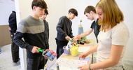 Zwei Personen stehen sich an einem Tisch gegenüber. Die linke Person malt mit einer Malerrolle auf einem vor ihr liegenden Stück Papier verschiedene Muster