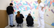 Drei Jugendliche stehen vor einer Wand und malen auf dieser bunte Dreiecke aus. Daneben steht eine Person in einem Maleroutfit