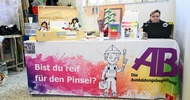Eine Person sitzt hinter einem Tisch und blickt auf einen Laptop. Auf dem Tisch ist ein Banner, auf dem Bist du reif für den Pinsel steht?