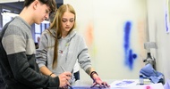 Zwei Personen stehen an einem Tisch und malen mit einer Sprühvorrichtung eine Schablone aus, die vor ihnen liegt