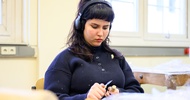 Eine Person mit Kopfhörern sitzt hinter einem Tisch und schnitzt an einem Stück Holz herum
