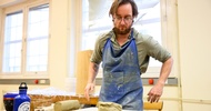 Eine Person mit Schürze steht hinter einem Tisch. Auf dem Tisch ist ein Klumpen Ton