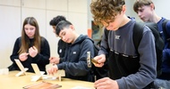 Eine jugendliche Person steht an einem Tisch und schlägt mit einem Hammer einen Nagel in ein flaches, kleines Holzstück