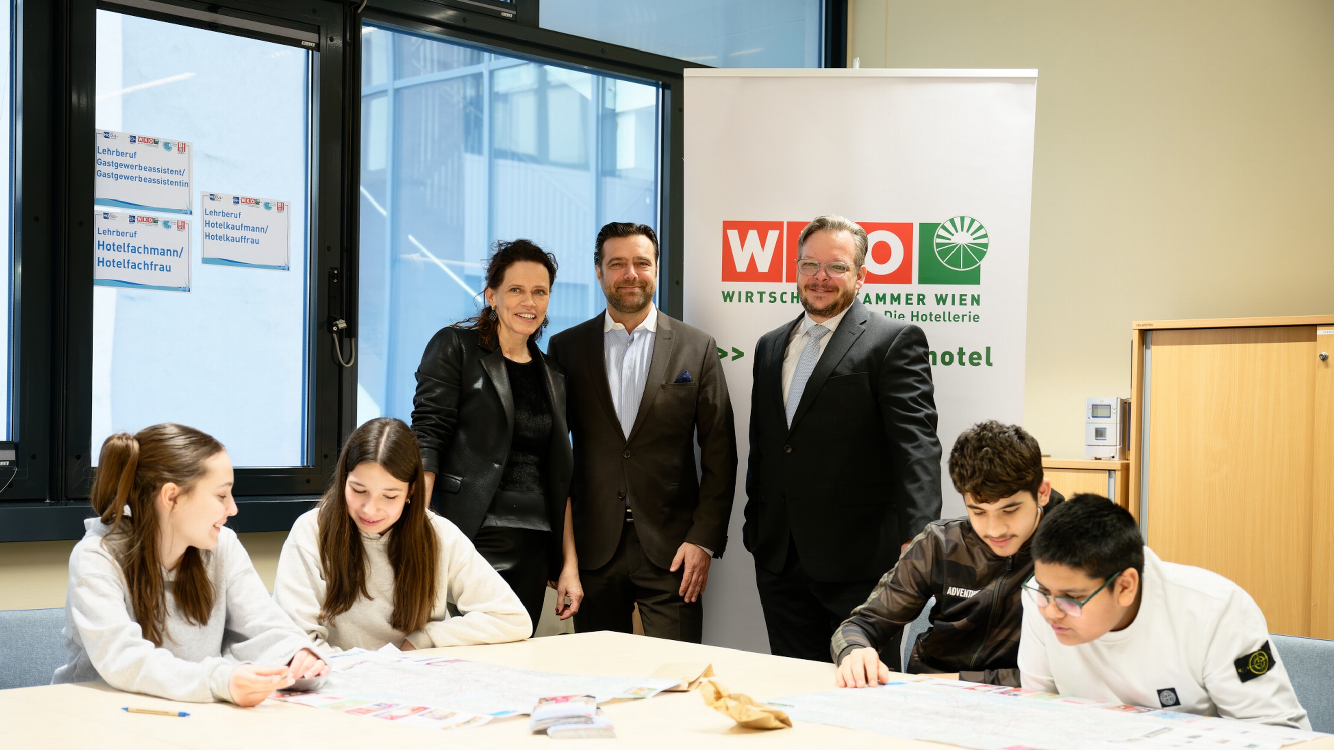 An einem Tisch sitzen vier Jugendliche in zwei Gruppen jeweils zu zweit. Beide Gruppen halten Stifte und schreiben Zettel. Am Tisch stehen nebeneinander drei Personen, die direkt in die Kamera blicken