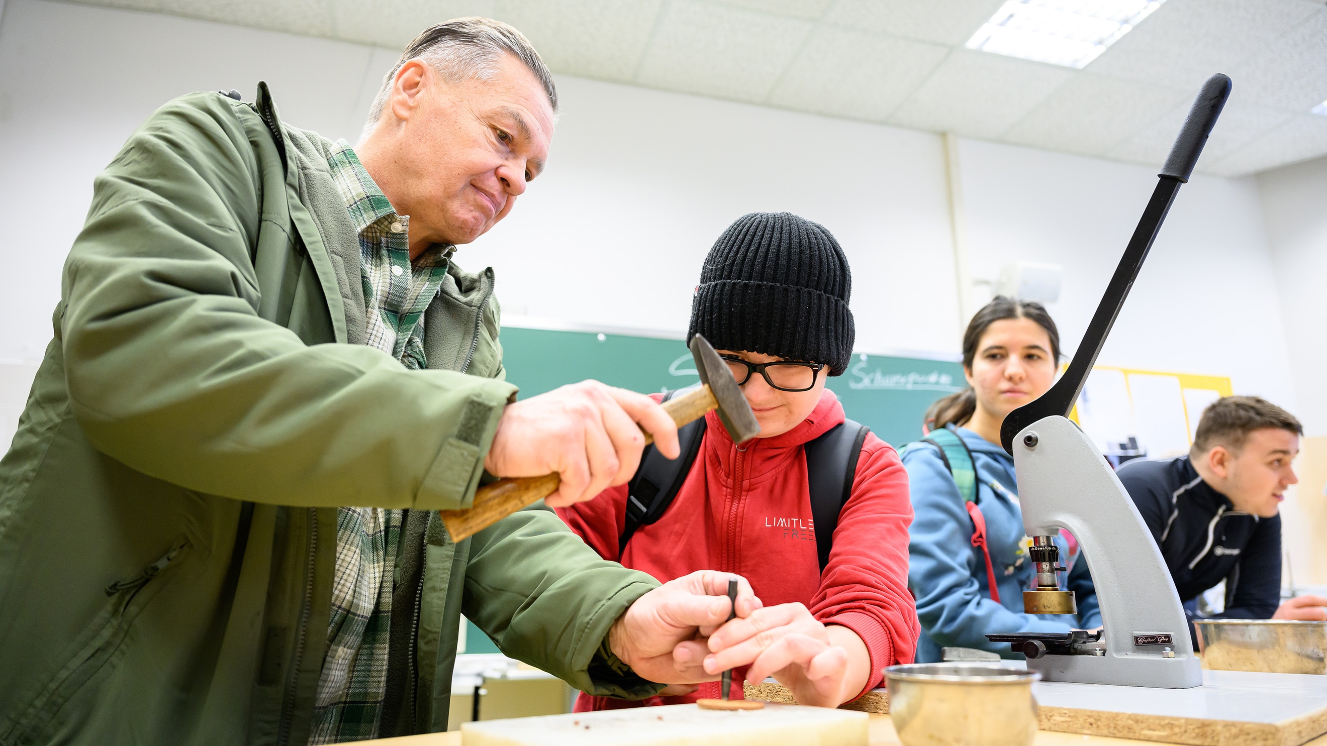 Eine Person und ein Kind stehen nebeneinander an einem Tisch. Die Person hilft dem Kind einen Nagel in ein flaches, rundes, kleines Holzstück zu schlagen