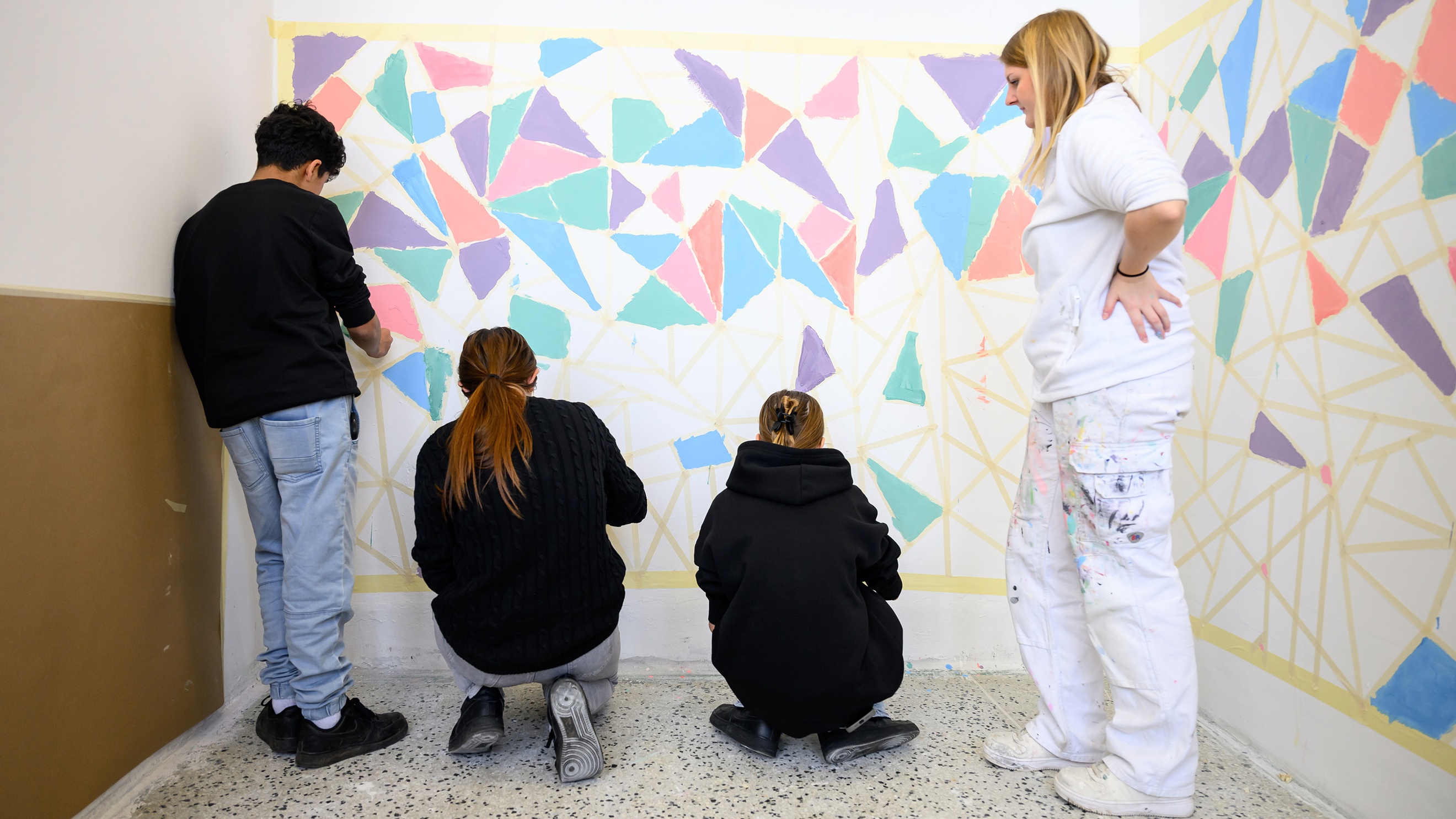 Drei Jugendliche stehen vor einer Wand und malen auf dieser bunte Dreiecke aus. Daneben steht eine Person in einem Maleroutfit