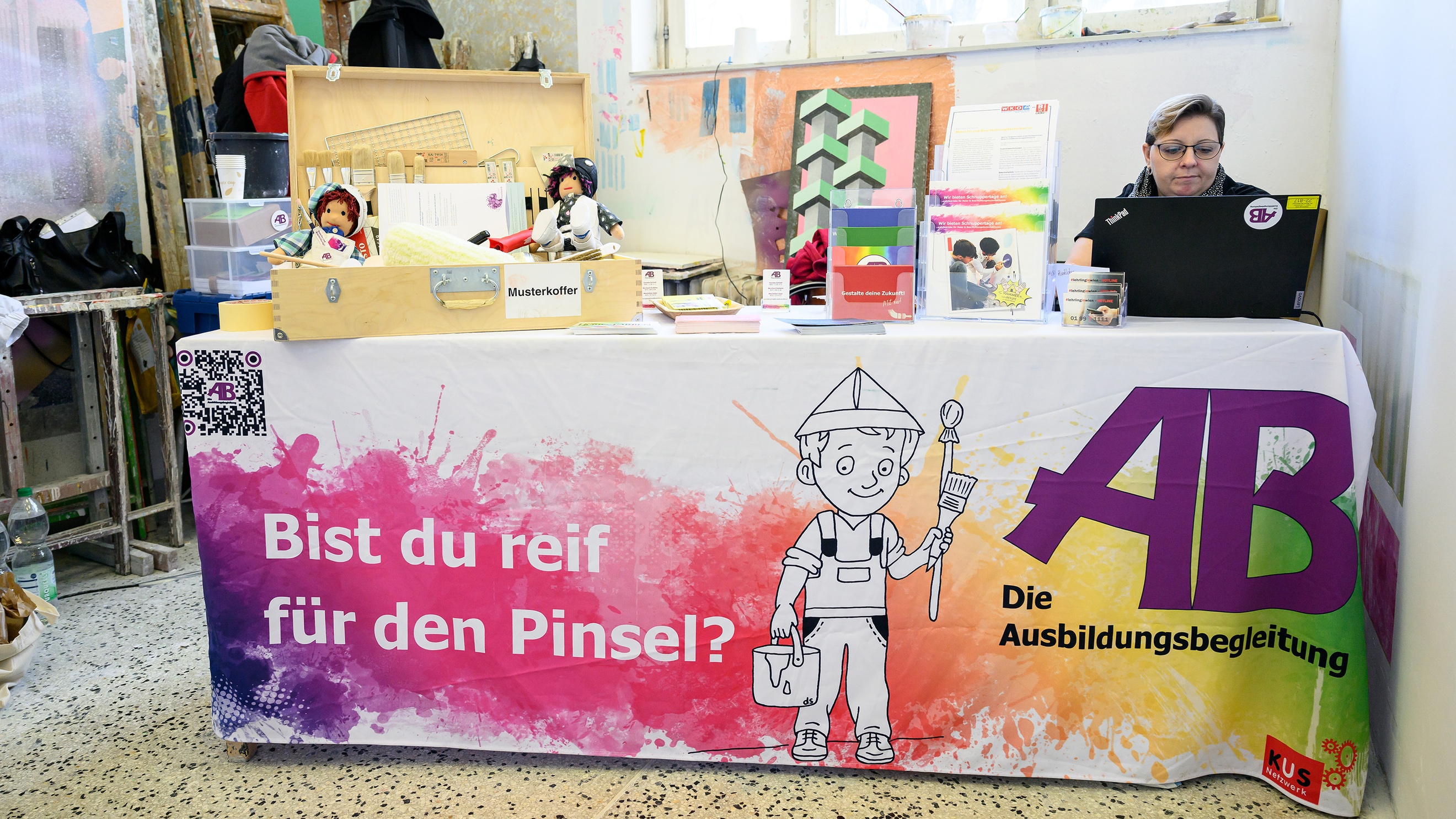 Eine Person sitzt hinter einem Tisch und blickt auf einen Laptop. Auf dem Tisch ist ein Banner, auf dem Bist du reif für den Pinsel steht?