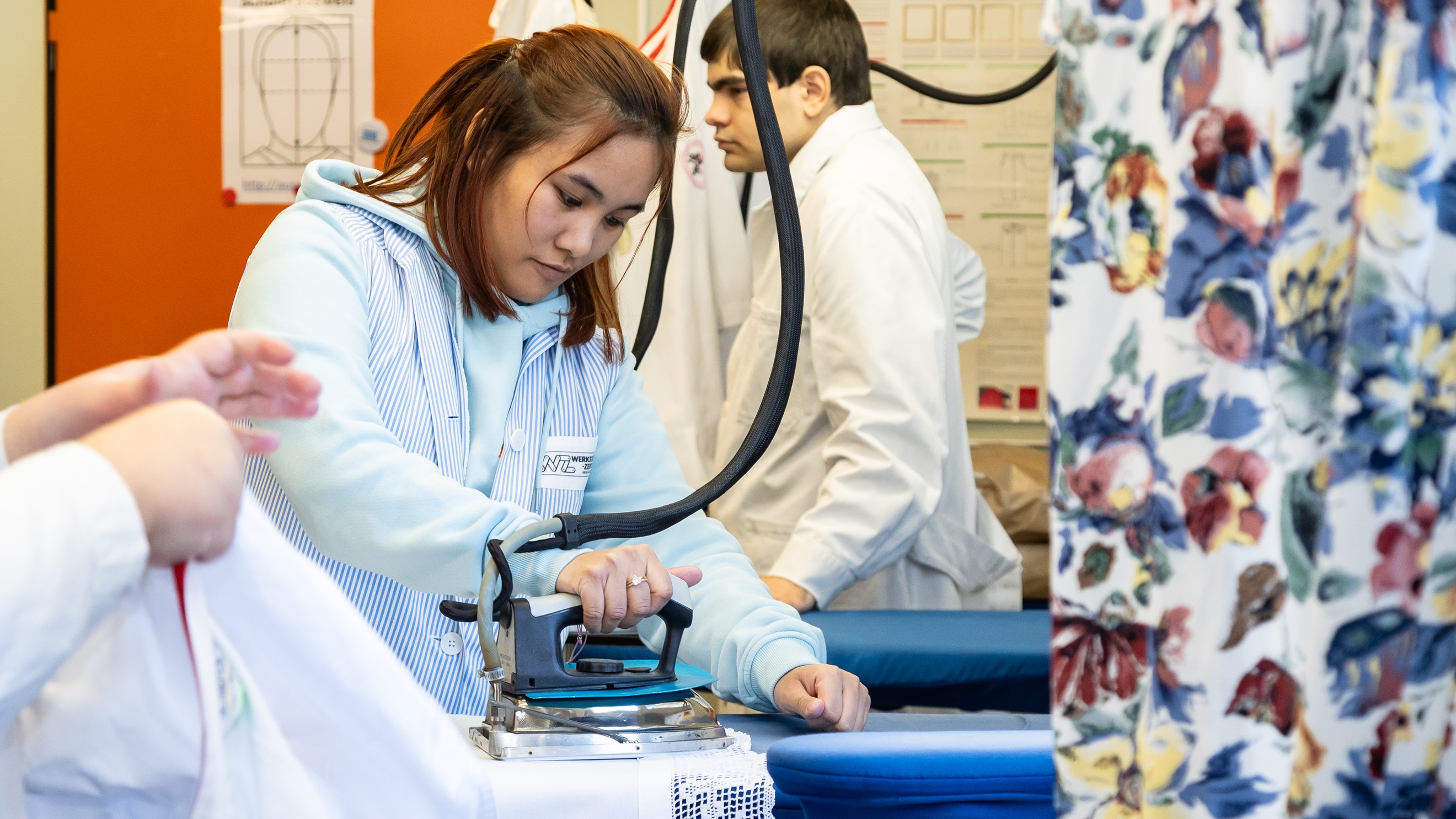 Seitliche Aufnahme einer Person, die ein helles Tischtuch auf einem Bügelbrett bügelt. Neben ihr stehen zwei weitere Personen neben Bügelbrettern