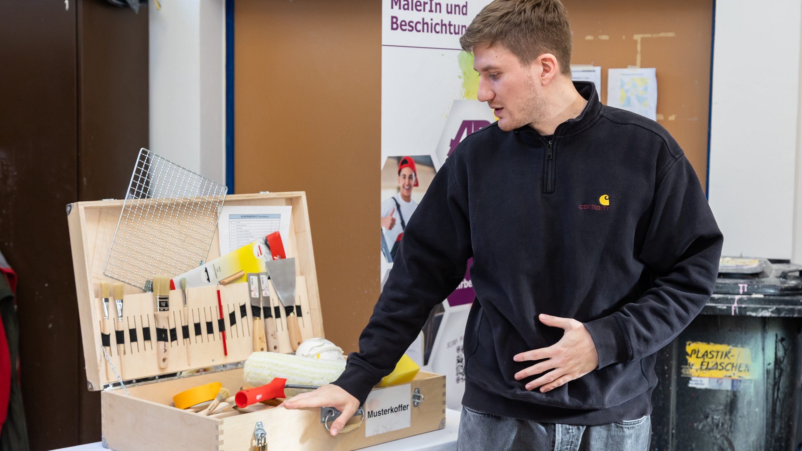 Eine Person mit leichtem Bart und kurzen Haaren steht an einem Tisch. Auf dem Tisch steht ein Holzkoffer, auf dem Musterkoffer steht. Im Koffer ist verschiedenes Werkzeug