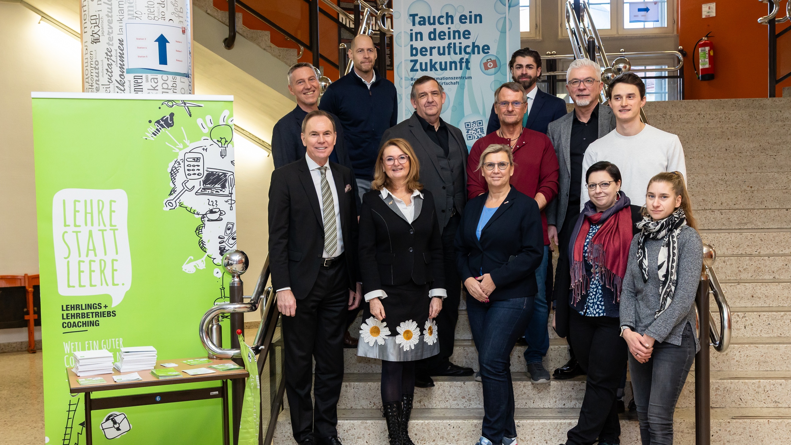 Porträt von mehreren Personen, die auf einer Treppe stehen. Hinter ihnen ist ein großer Banner, auf dem Tauch ein in deine berufliche Zukunft steht. Links neben ihnen ist ein weiterer Banner, auf dem Lehre statt Leere steht