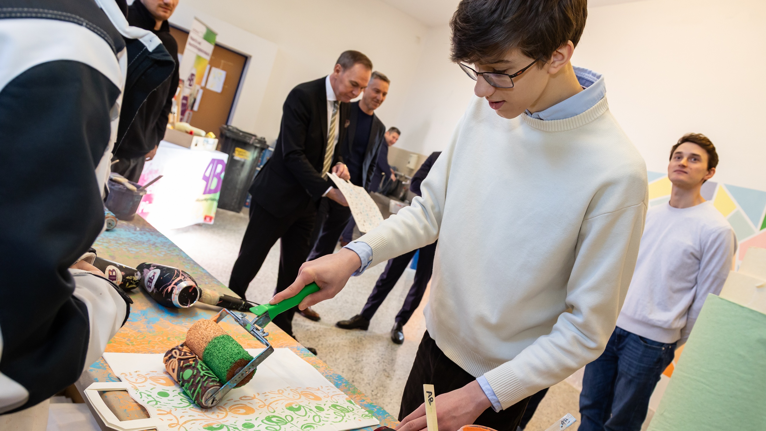Eine jugendliche Person mit Brille und kurzen Haaren steht an einem Tisch und malt mit einer Malerrolle Muster auf ein weißes Papiersackerl. Im Hintergrund stehen mehrere Personen