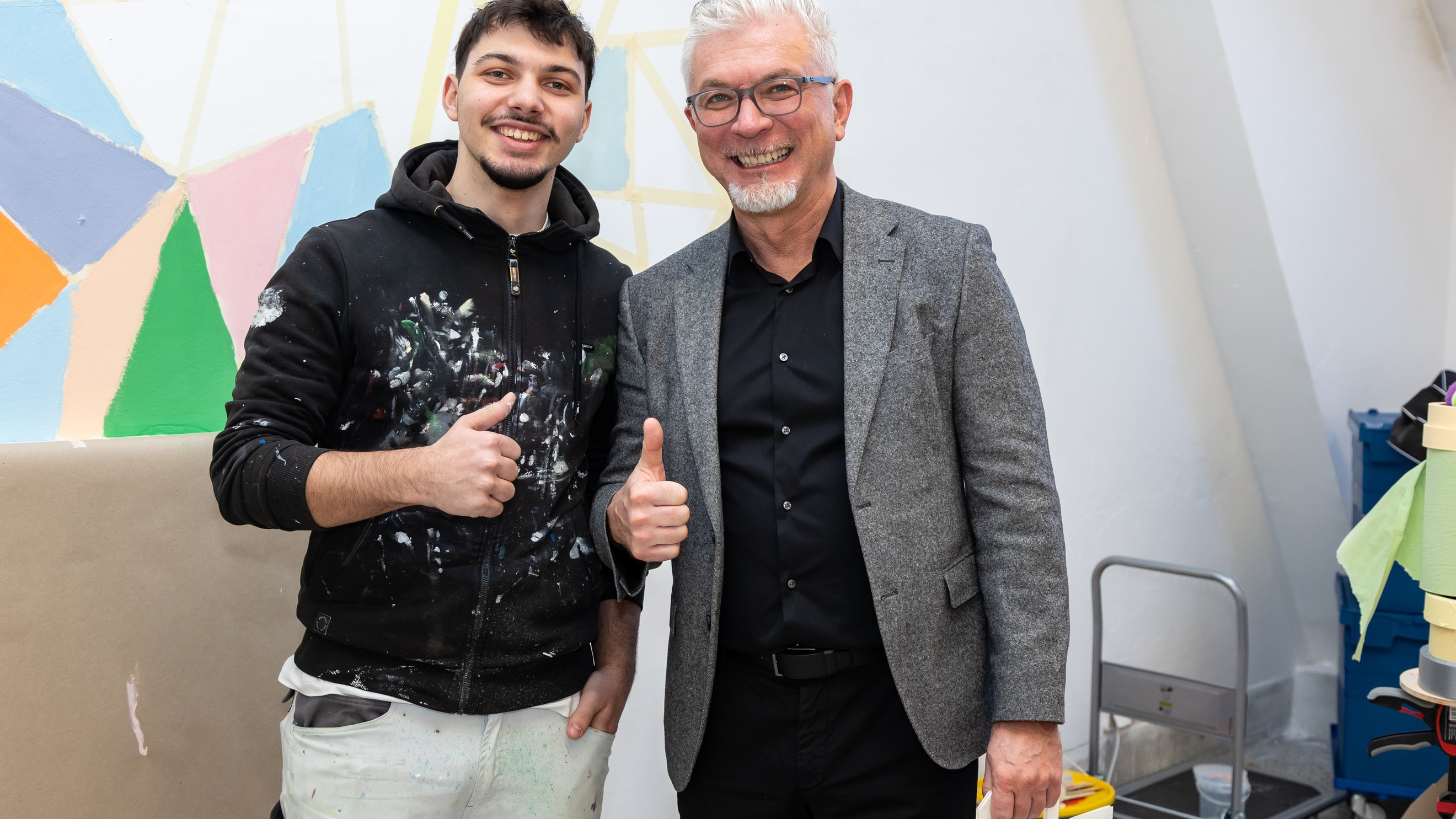 Porträt von zwei lächelnden Personen, die vor einer teilweise bemalten Wand stehen und den Daumen hoch halten. Auf der Kleidung der linken Person sind Farbkleckser