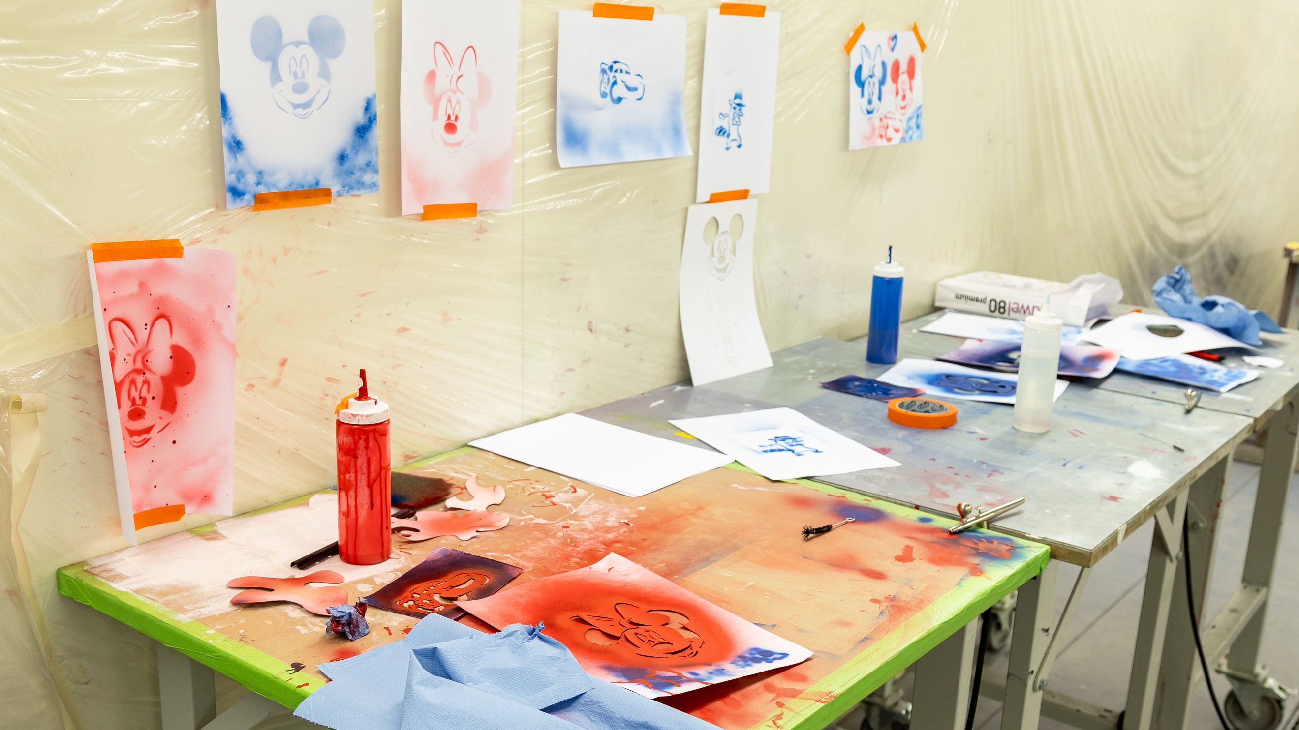 Auf drei nebeneinander stehenden Tischen liegen Zettel, in denen die Köpfe von Comicfiguren ausgeschnitten und bunt bemalt sind. An den Wänden hängen Zettel mit den Zeichnungen