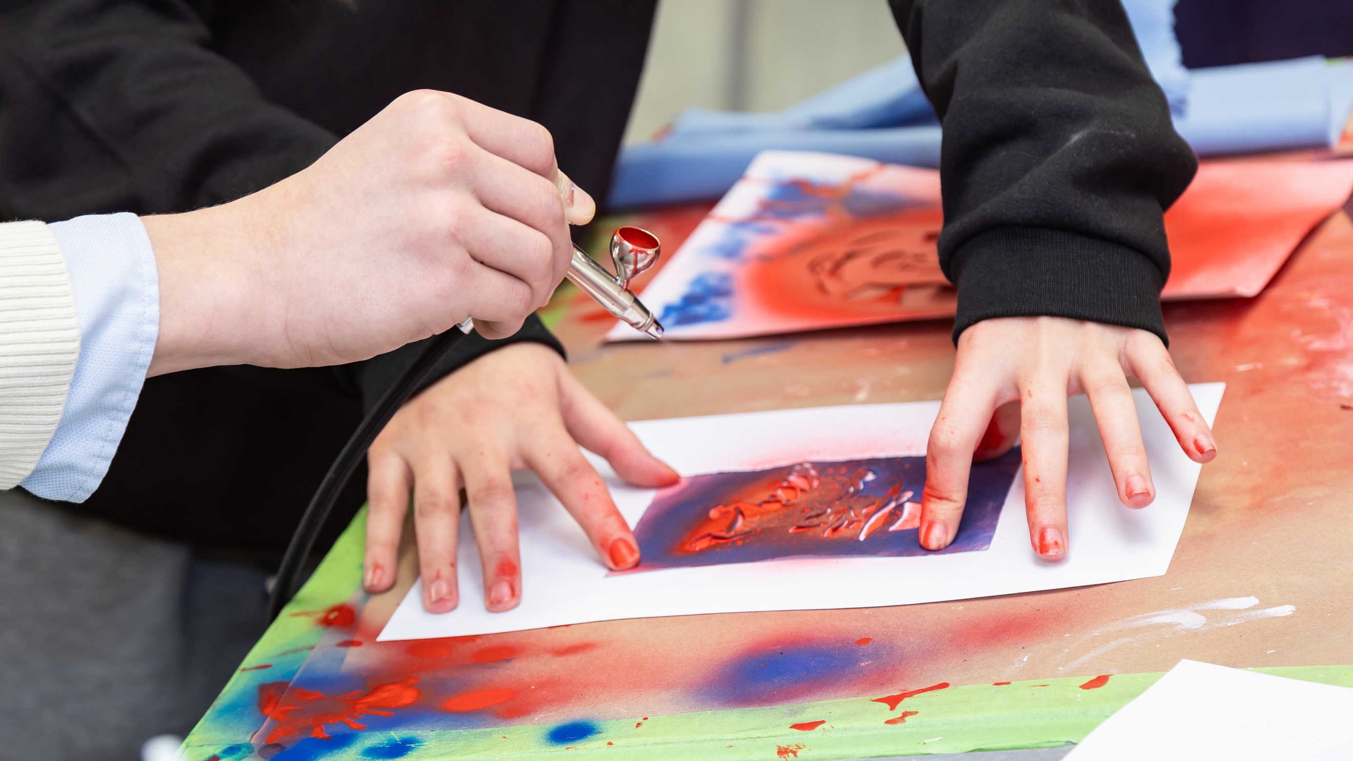 Nahaufnahme der Hände einer Person, die einen Zettel festhält, aus dem eine Figur ausgeschnitten ist. Eine andere Person hält eine kleine Farbpistole auf diesen Bereich