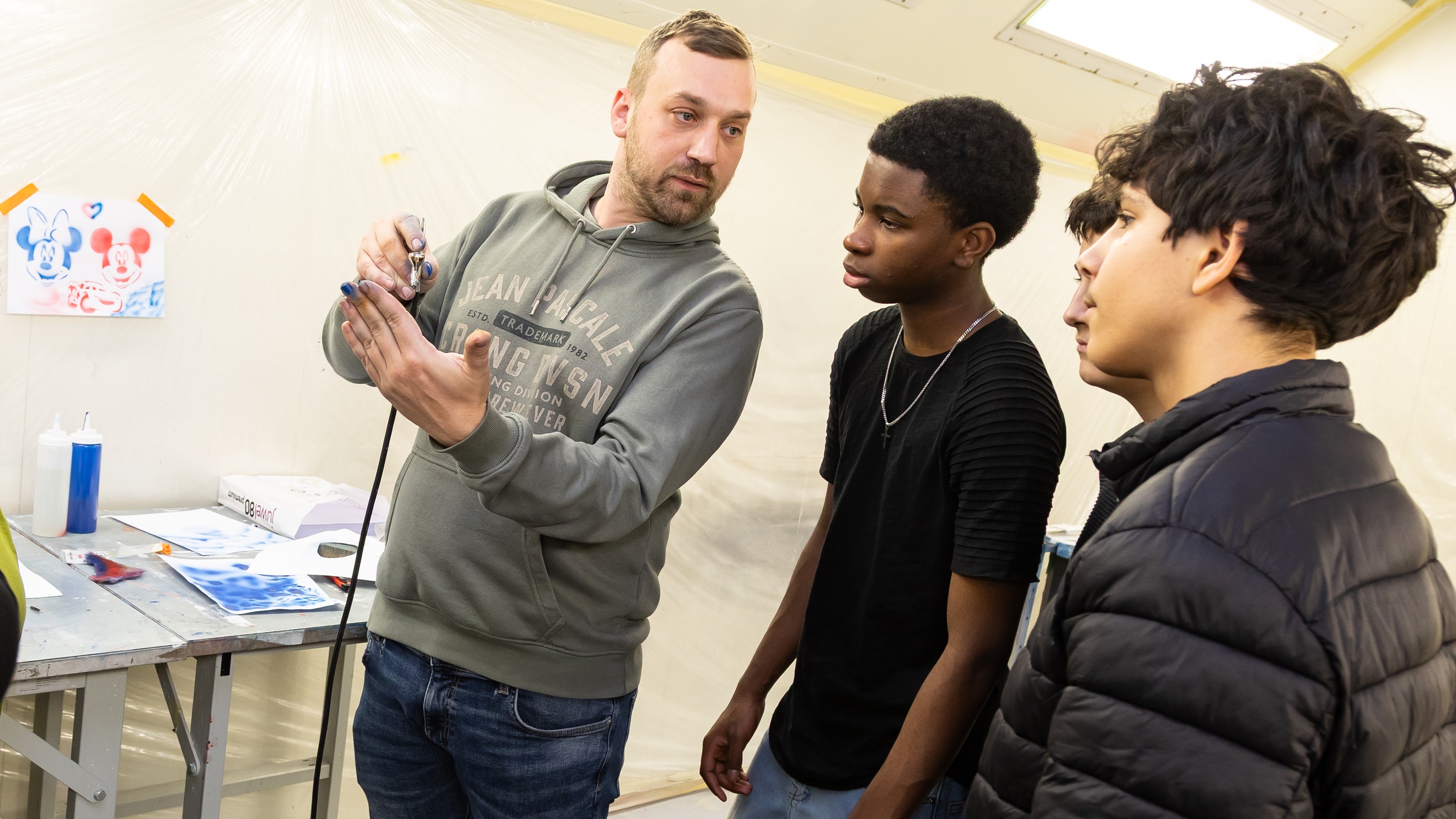 Eine Person mit Bart und kurzen Haaren steht neben einem Tisch und hält eine kleine Farbpistole auf ihre linke Hand. Neben ihr stehen drei jugendliche Personen, die auf die Hand der Person blicken