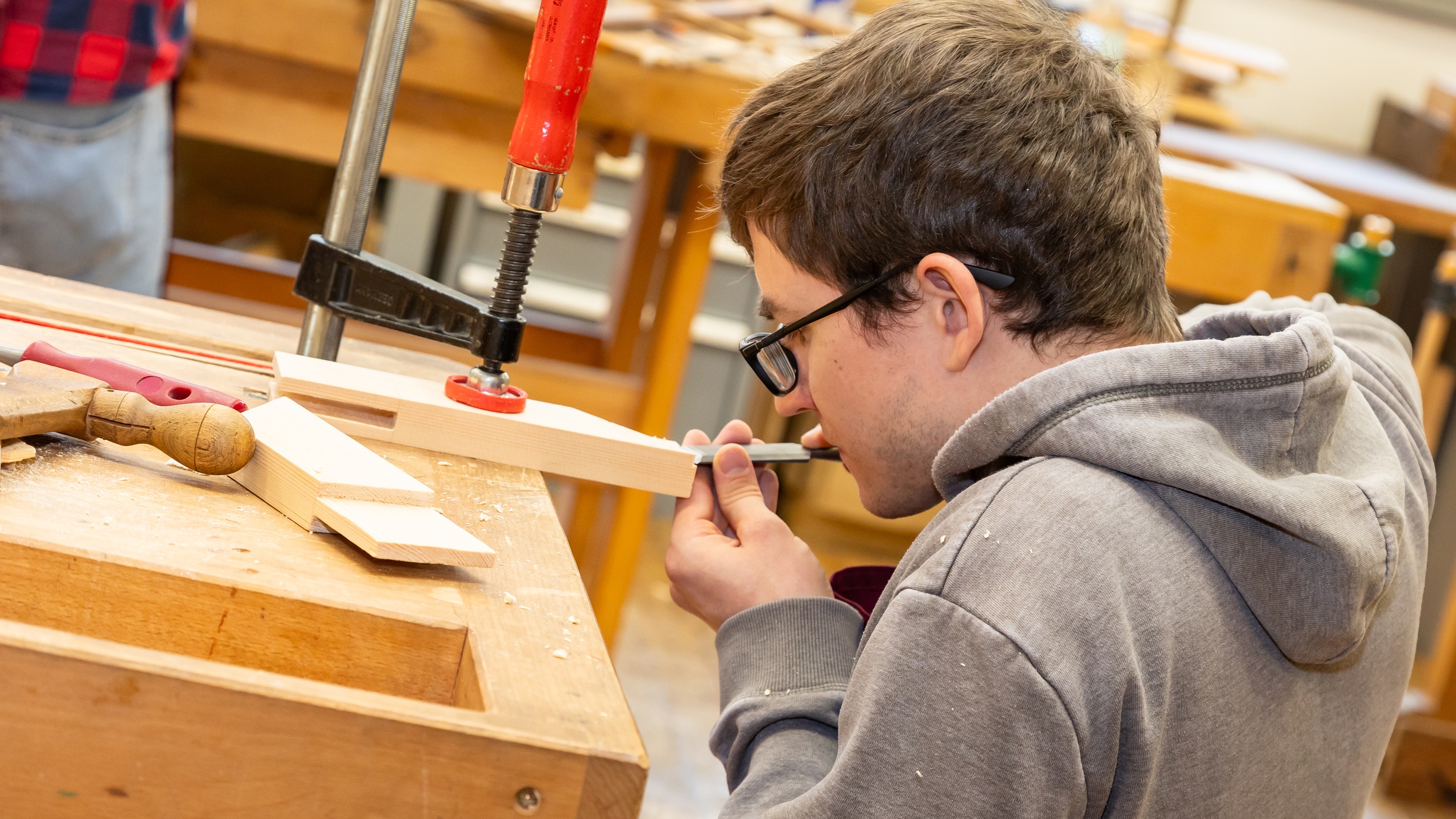 Seitliche Aufnahme einer Person mit Brille, die neben einem Werktisch hockt und mit einem Werkzeug ein kleines Holzstück bearbeitet, das eingespannt ist