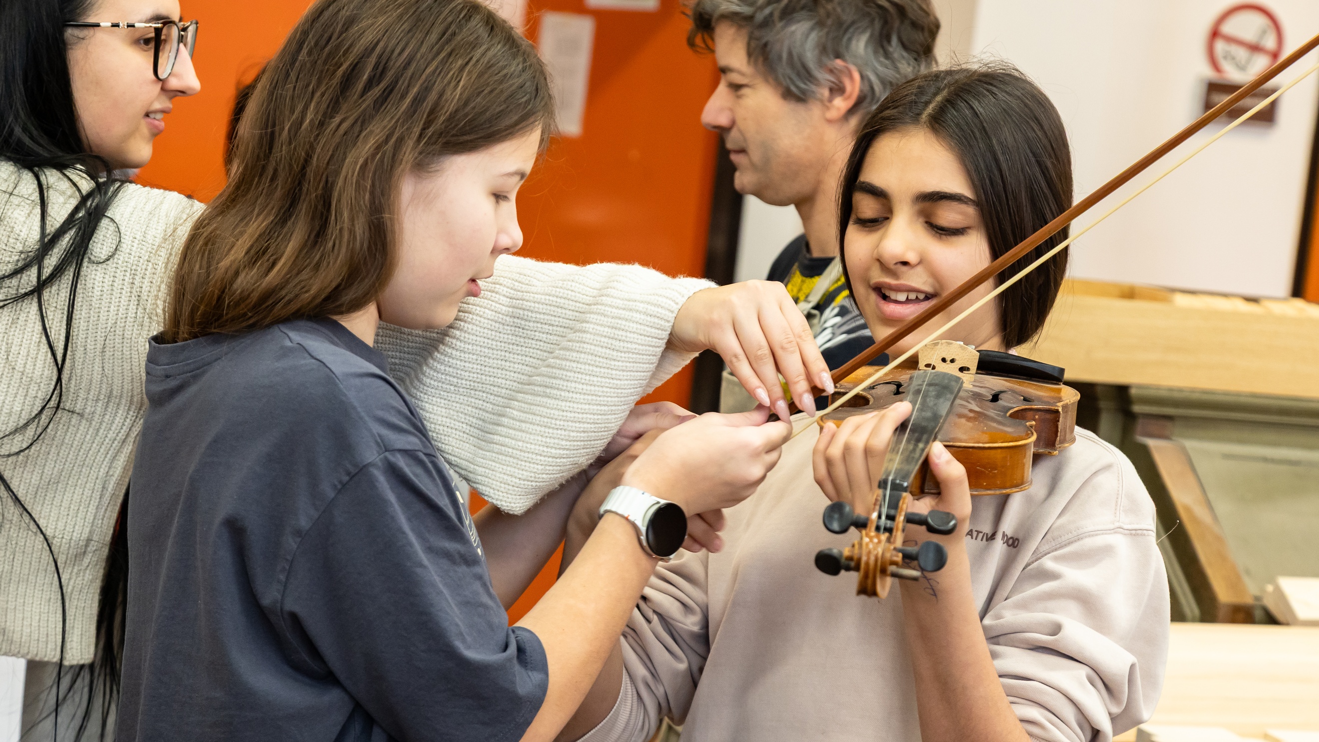 Eine jugendliche Person hält eine Geige. Neben ihr stehen zwei Personen, die den Bogen zur Geige halten