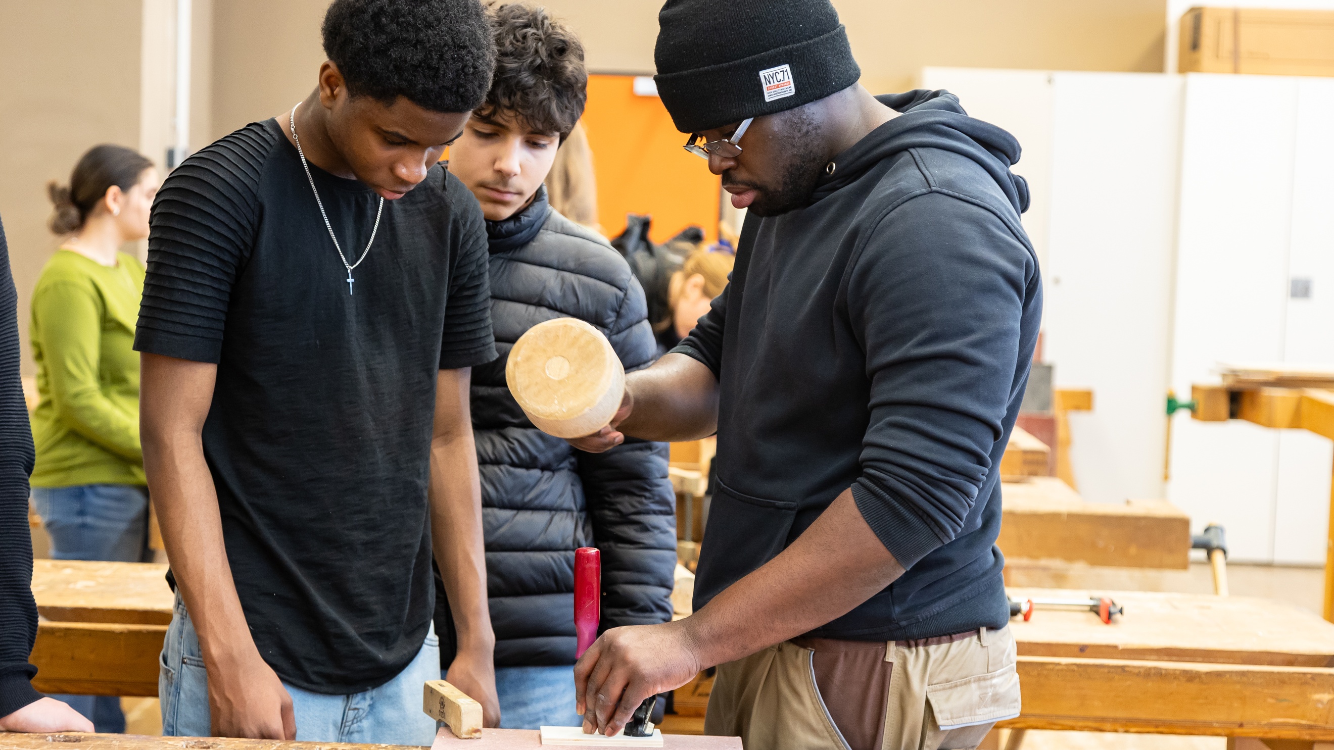 An einem Werktisch steht eine Person, die auf ein eingespanntes Stück Holz einen Schraubenzieher hält. In der anderen Hand hält sie einen runden Hammer. Zwei jugendliche Personen blicken auf den Schraubenzieher