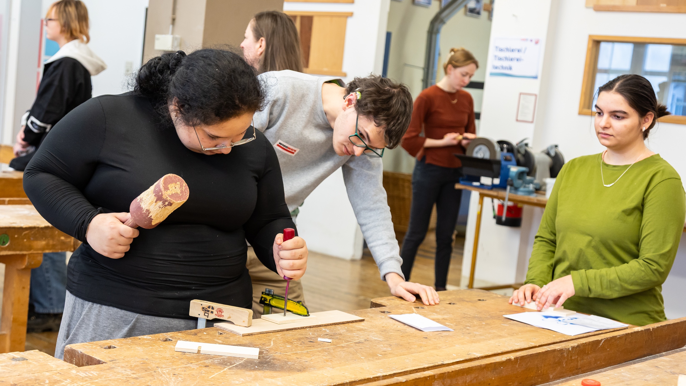 An einem Werktisch steht eine jugendliche Person, die ein dünnes, eingespanntes Holzbrett mit einem Schraubenzieher bearbeitet. Eine andere Person steht direkt neben ihr und blickt auf das Holzstück
