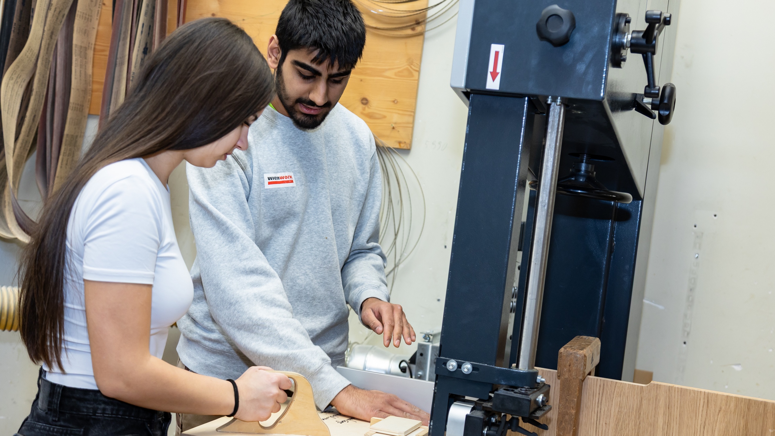 Seitliche Aufnahme zweier jugendlicher Personen, die an einer Säge stehen und Holzstücke auseinander schneiden