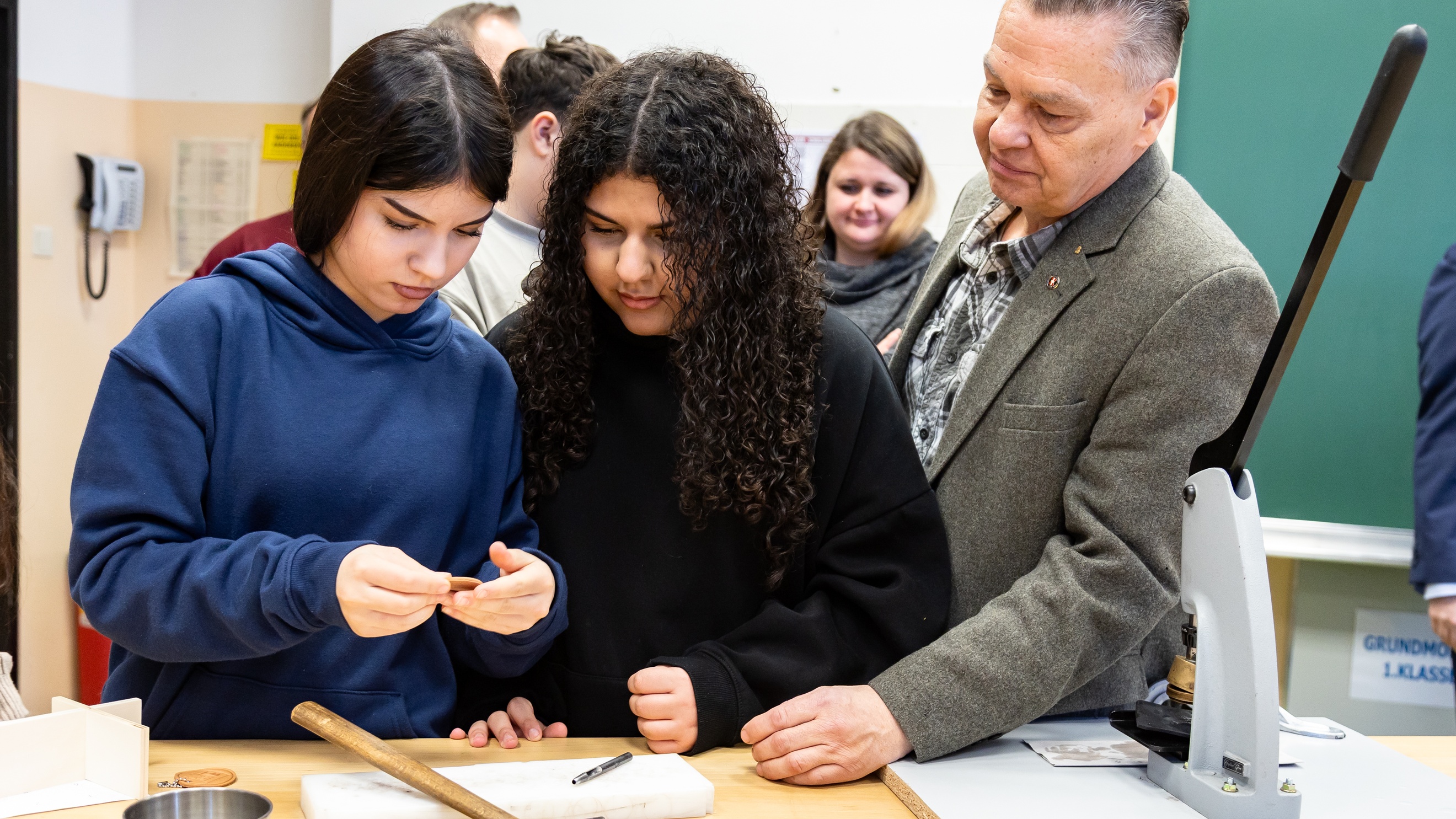 Zwei jugendliche Personen stehen an einem Werktisch. Die linke Person hält ein kleines Holzstück, auf das beide blicken. Auf dem Tisch liegt ein Hammer. Eine weitere Person steht neben ihnen