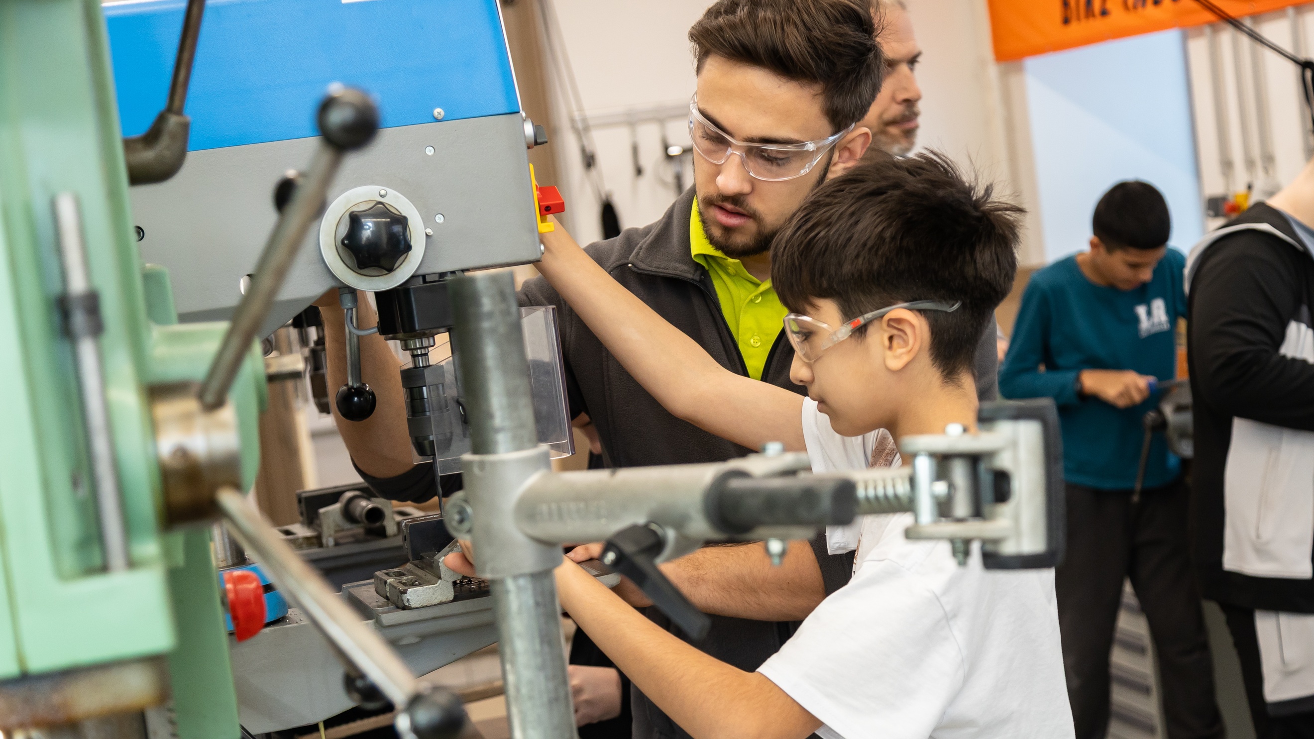 Eine Person mit Schutzbrille und ein Kind mit Schutzbrille stehen nebeneinander an einem Gerät in einer Werkstatt