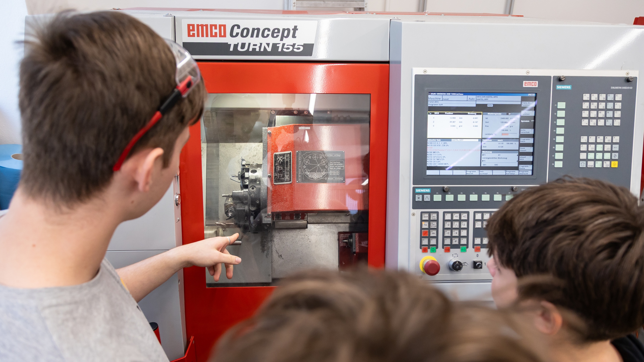 Rückenansicht von drei Personen, die nebeneinander ovr einem Gerät mit Schaltflächen steht. Auf dem Gerät steht Emco Konzept