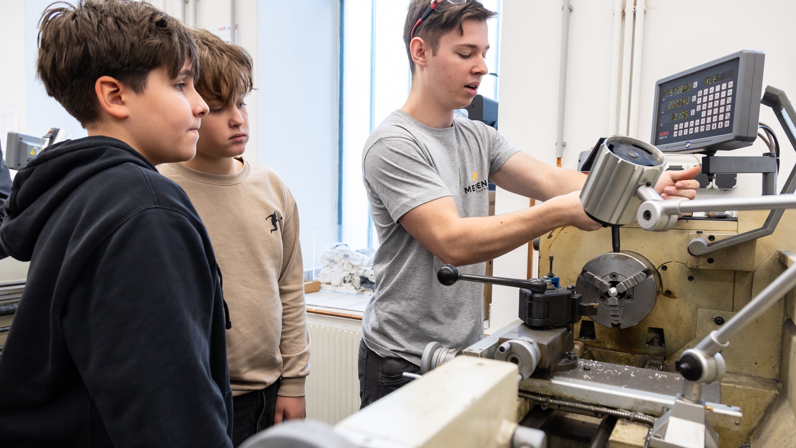 Zwei Kinder und eine jugendliche Person mit Schutzbrille im Haar stehen neben einem sehr großen Werkzeug. Die jugendliche Person spricht und bedient das Werkzeug
