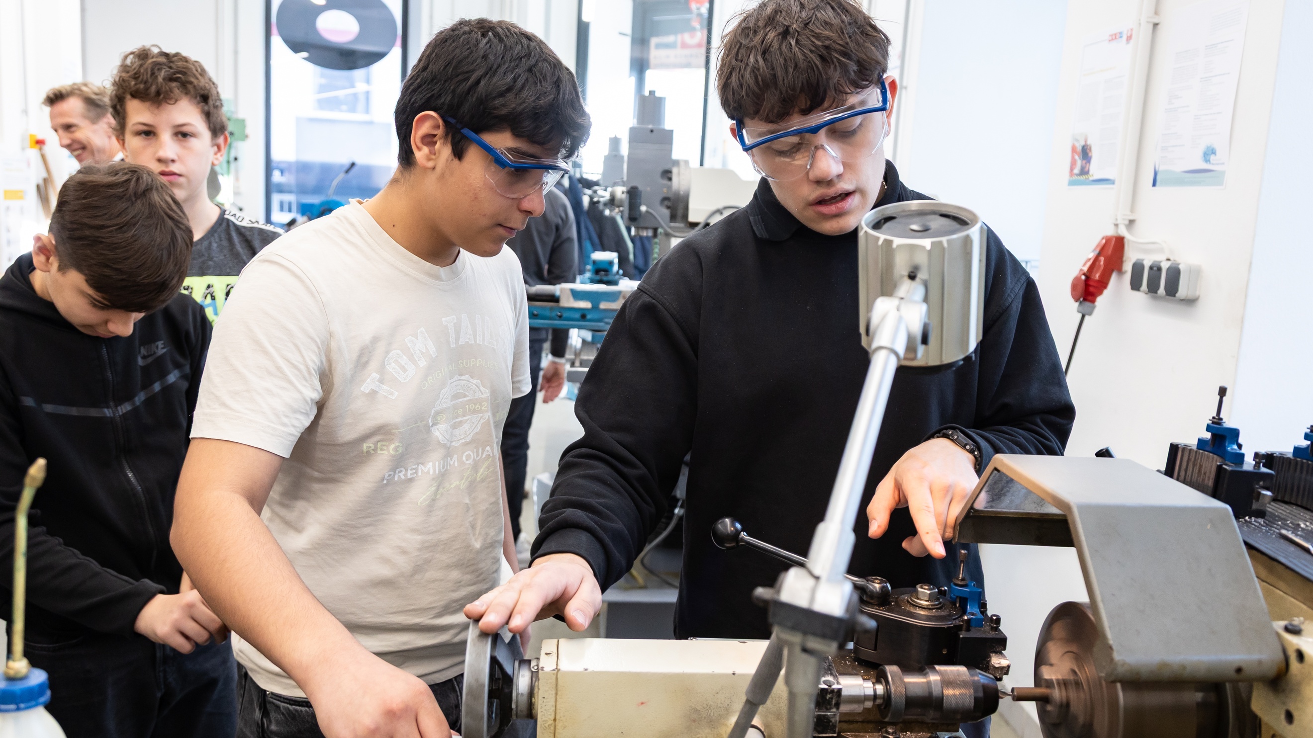 Zwei jugendliche Personen mit Schutzbrillen stehen an einem Gerät zur Bearbeitung von Metall