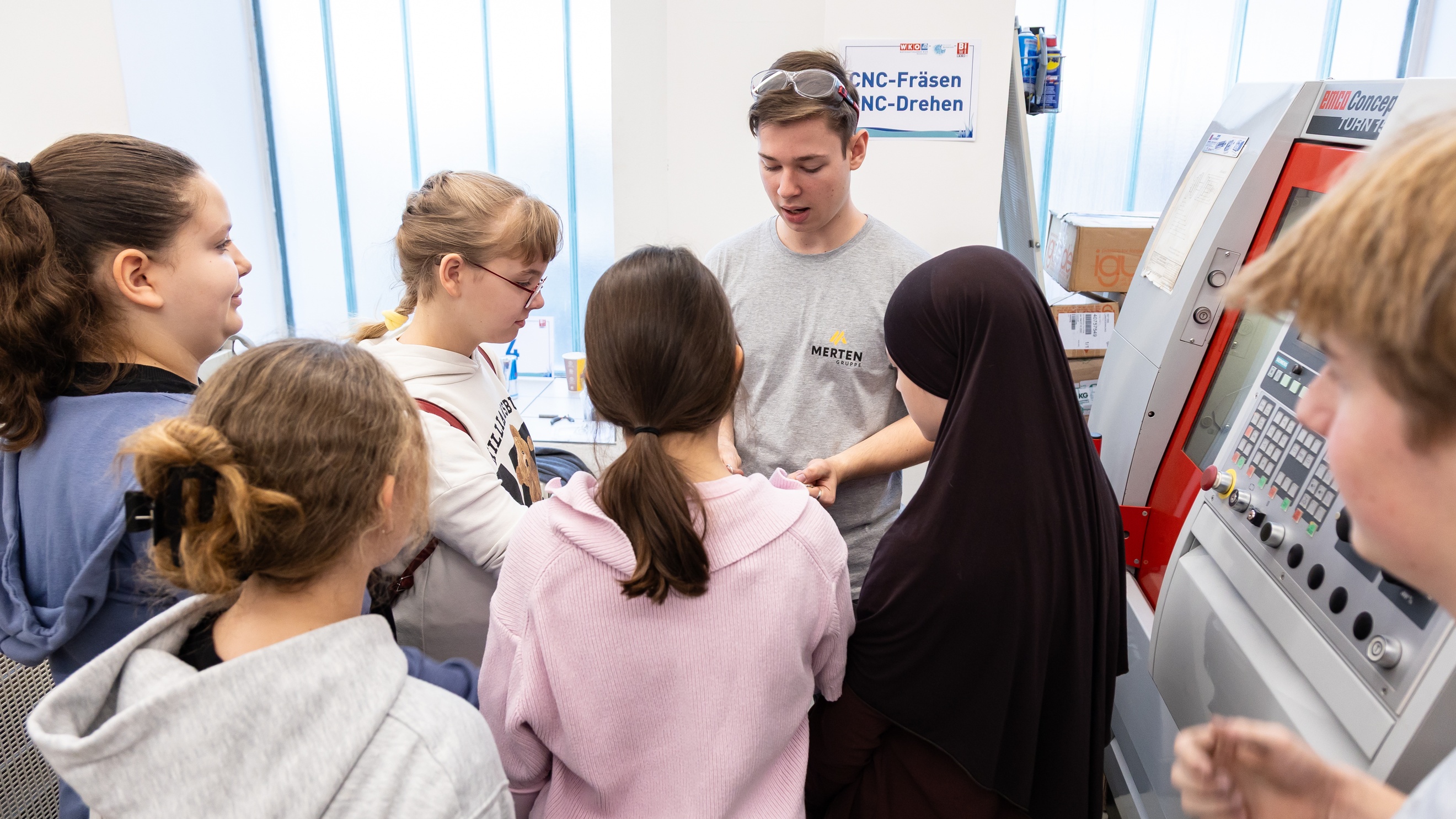 Sechs Kinder stehen neben einer jugendlichen Person mit Schutzbrille. Neben ihnen steht ein Gerät
