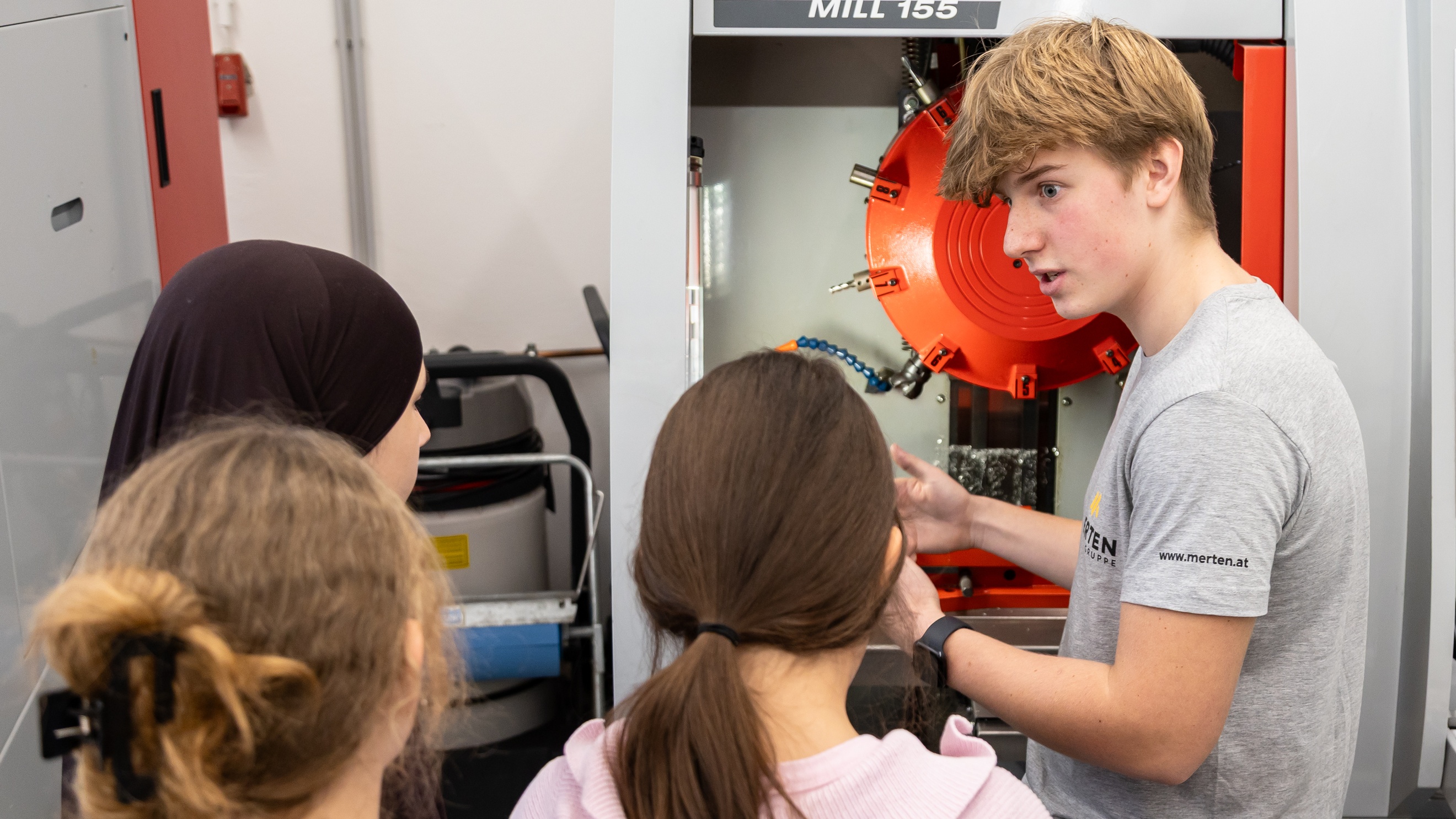 Drei Kinder stehen neben einer jugendlichen Person mit Schutzbrille. Neben ihnen steht ein Gerät