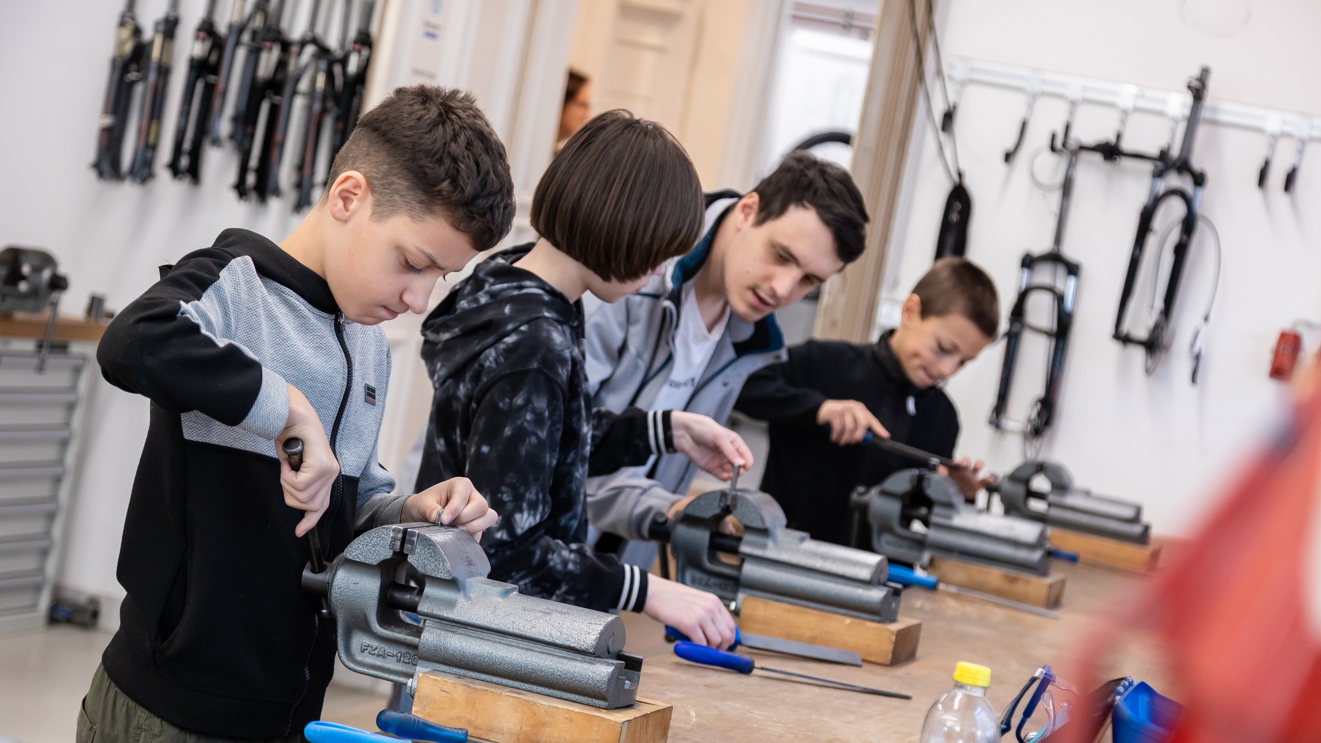 Drei Jugendliche stehen nebeneinander an einem Tisch mit Drehbänken und probieren diese aus. Neben einem Kind steht eine erwachsene Person, die ihm etwas erklärt<br />
