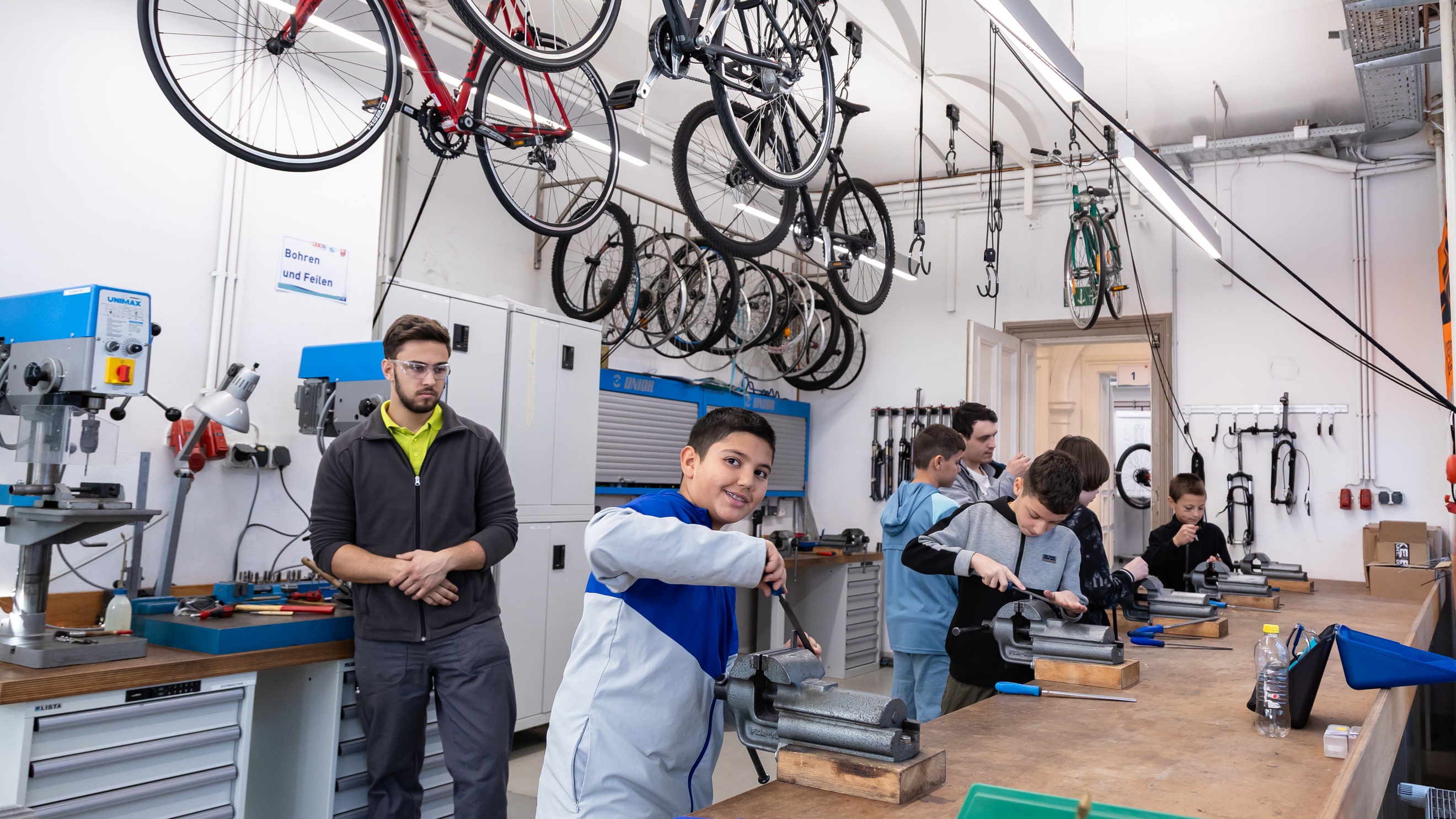 In einem großen Raum stehen vier jugendliche an einem Tisch mit Drehbänken und probieren diese aus. Eine Person mit Schutzbrille steht im Hintergrund. Von der Decke hängen Fahrräder<br />
<br />

