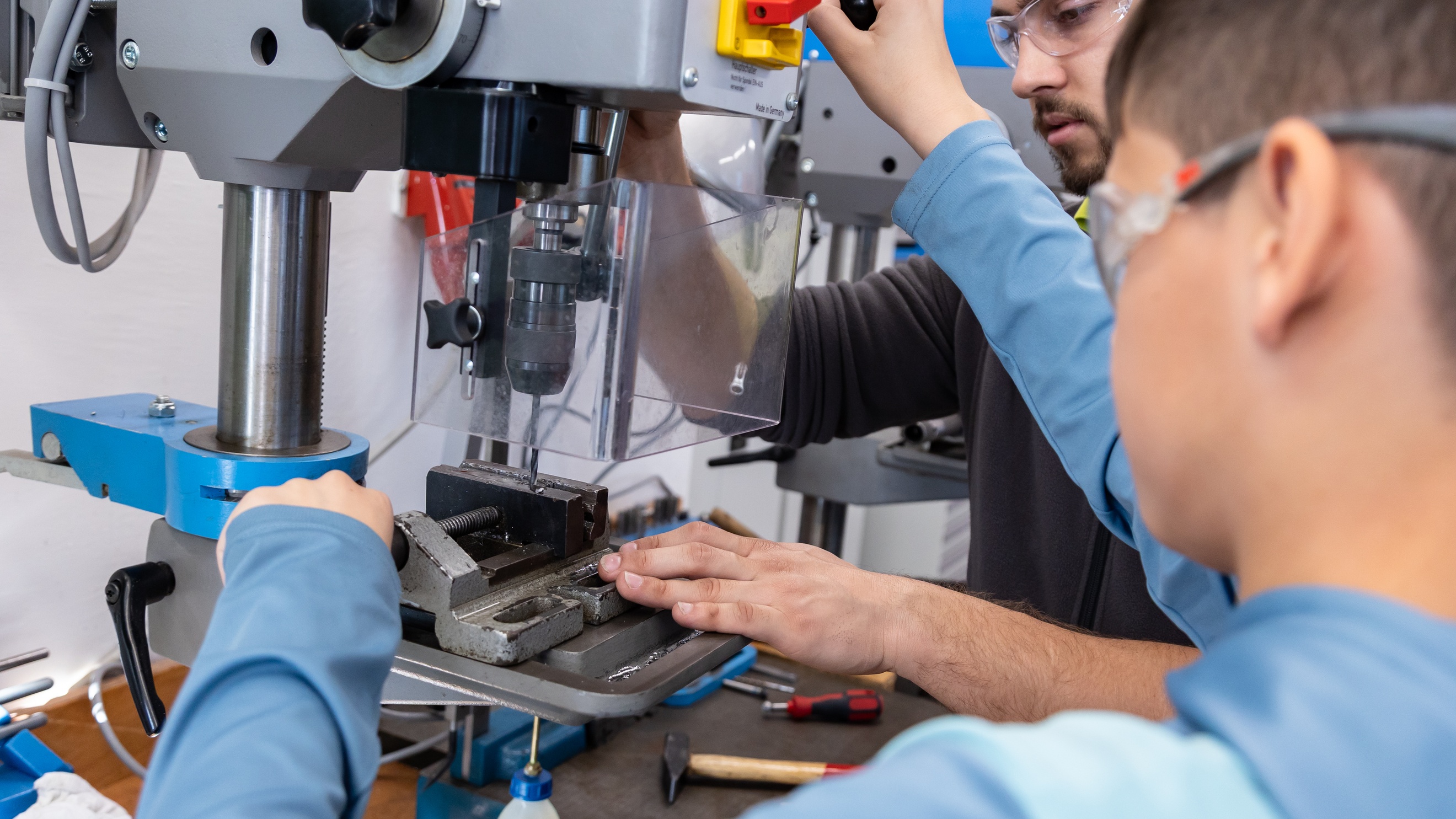Eine jugendliche Person mit Schutzbrille bedient einen Metallbohrer. Eine erwachsene Person mit Schutzbrille steht neben ihr und hilft ihr den Bohrer zu bedienen