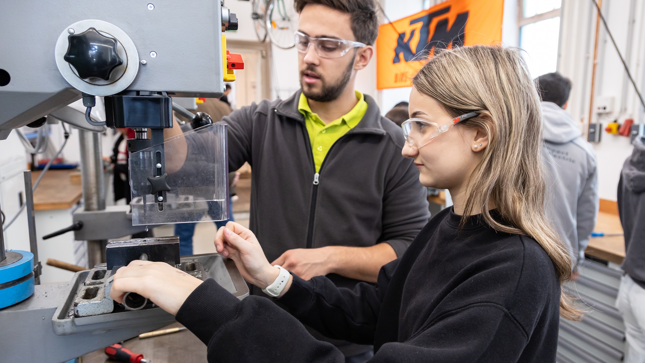 Eine jugendliche Person mit Schutzbrille bedient einen Metallbohrer. Eine erwachsene Person mit Schutzbrille steht neben ihr und hilft ihr den Bohrer zu bedienen