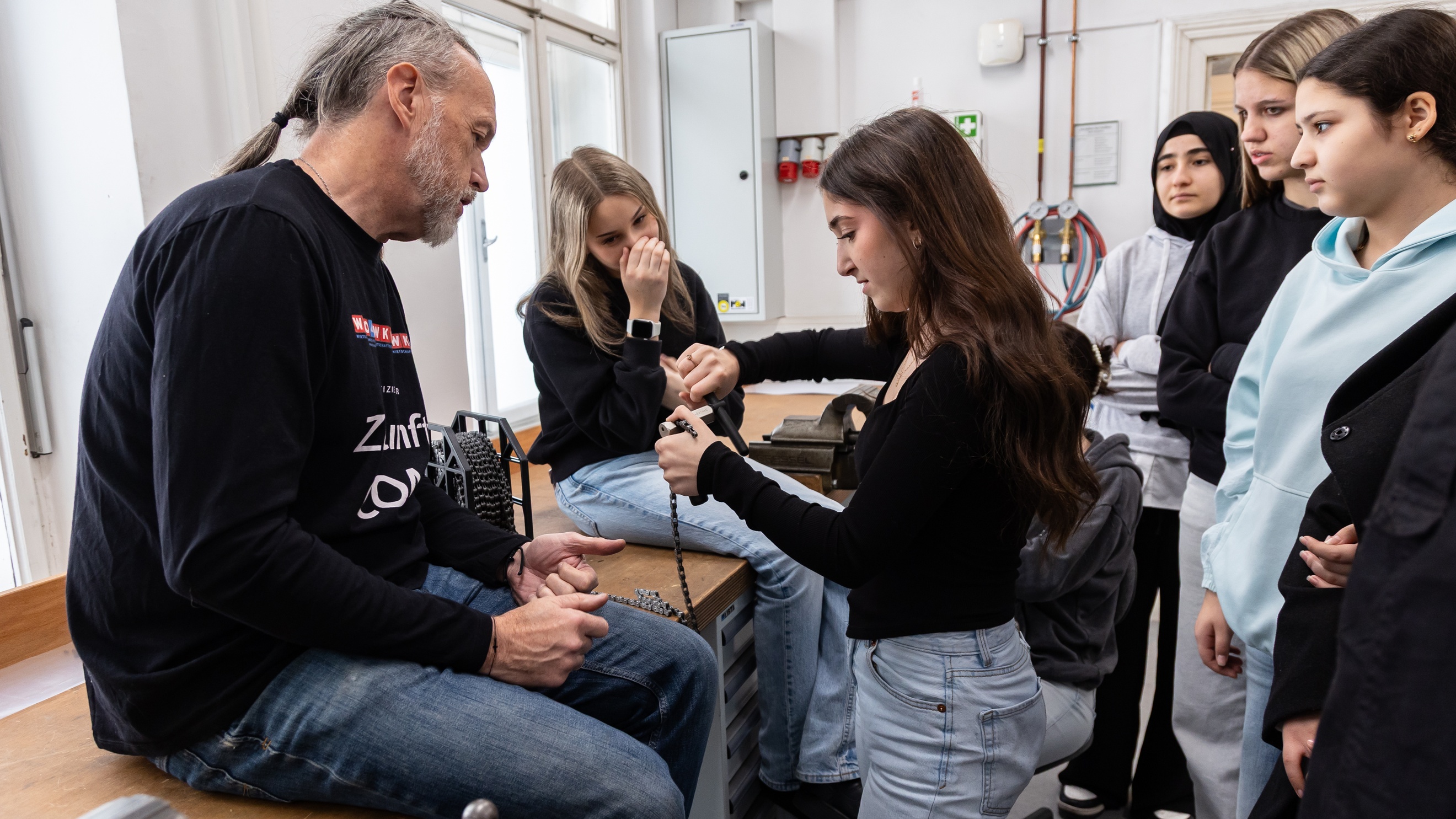 Eine jugendliche Person steht an einem Tisch und probiert ein Werkzeug aus. Eine erwachsene Person unterstützt sie. Weitere jugendliche Personen stehen daneben