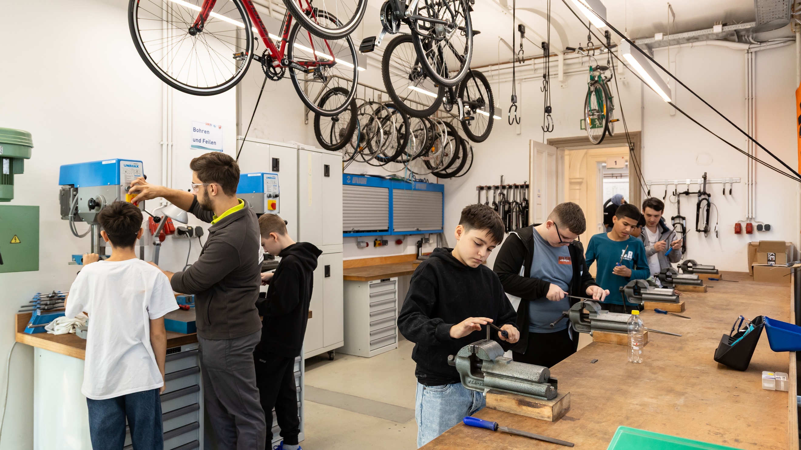 In einem großen Raum stehen vier jugendliche an einem Tisch mit Drehbänken und probieren diese aus. Zwei weitere Kinder und eine Person mit Schutzbrille stehen an einem Gerät. Von der Decke hängen Fahrräder
