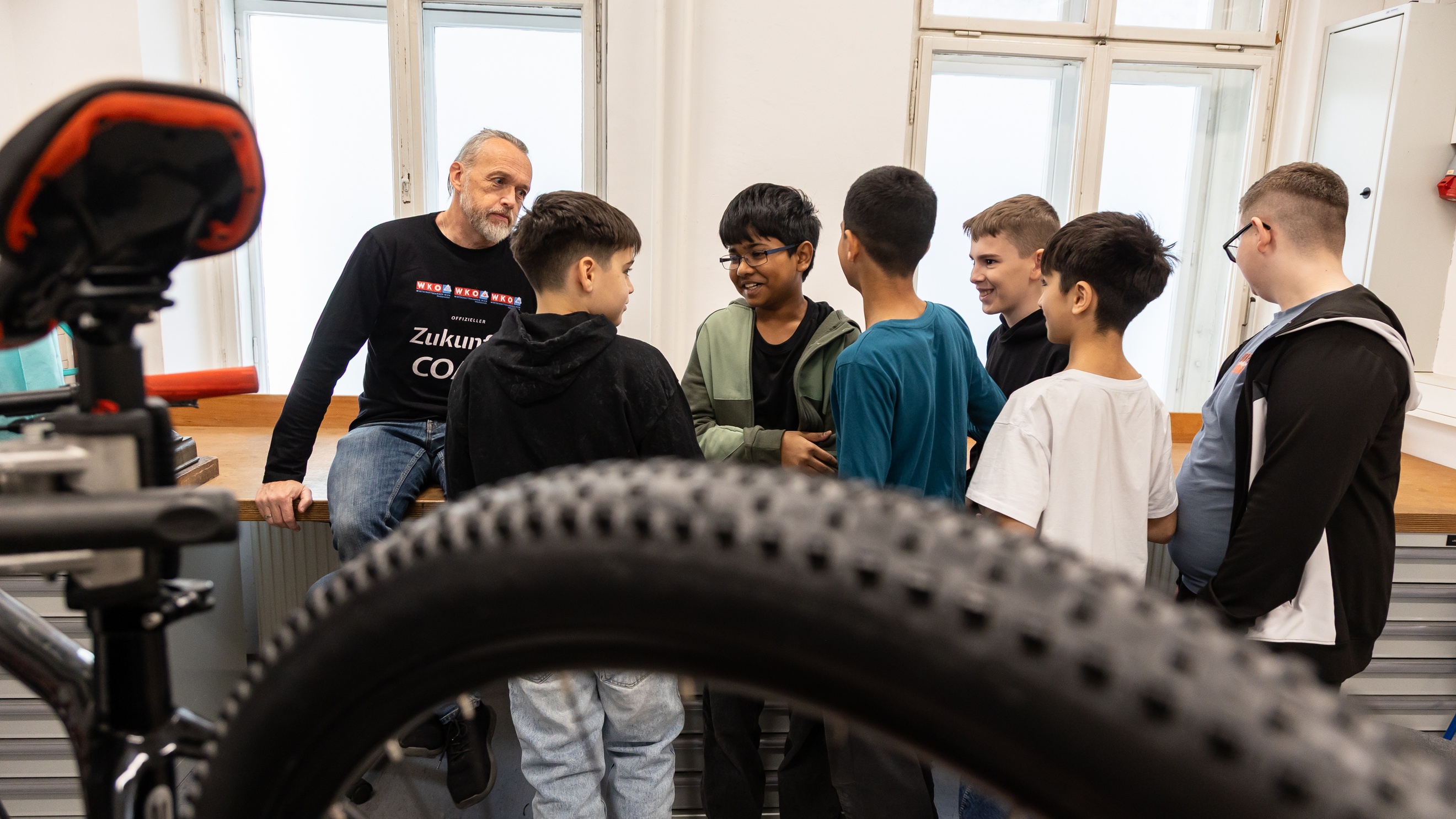 Im Vordergrund ist der hintere Teil eines Fahrrads. Dahinter stehen sechs Kinder in einem Kreis und unterhalten sich. Daneben steht eine Person