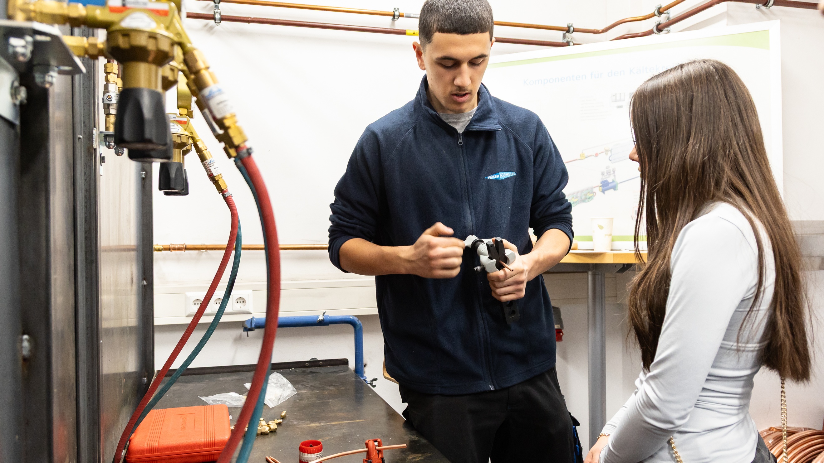 Eine Person mit einem Werkzeug in der Hand und eine jugendliche Person stehen nebeneinander neben einem Metalltisch. Auf dem Tisch liegen Kupferdrähte
