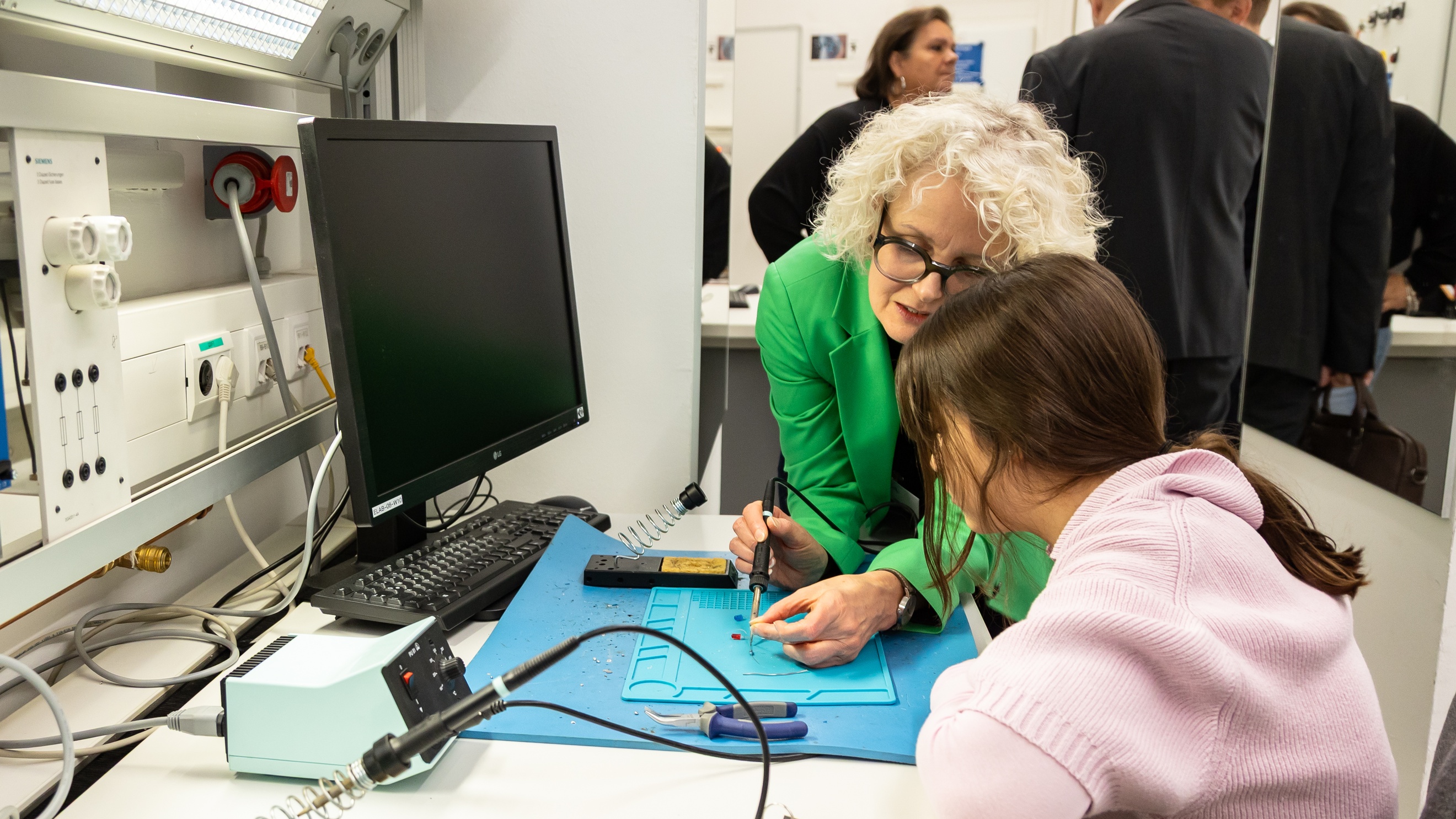 Ein Kind sitzt an einem Tisch. Neben ihr ist eine erwachsene Person, die einen Schraubenzieher mit einem Kabel hält. Auf dem Tisch liegen verschiedene Elektroden und Werkzeug