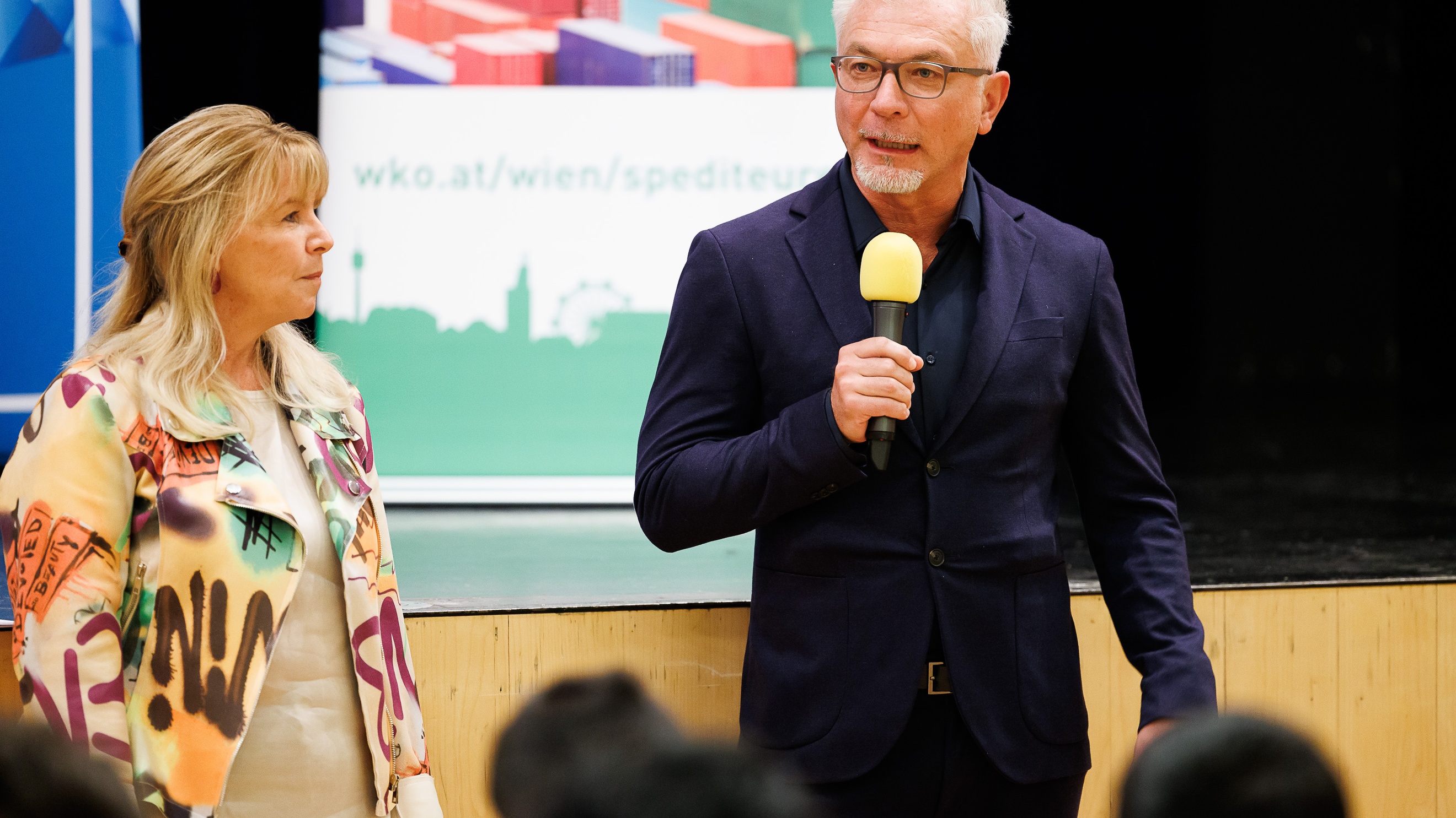 Frontalaufnahme zweier nebeneinander stehender Personen. Die rechte Person spricht in ein Mikrofon. Ihnen gegenüber sitzen mehrere Personen auf Stühlen
