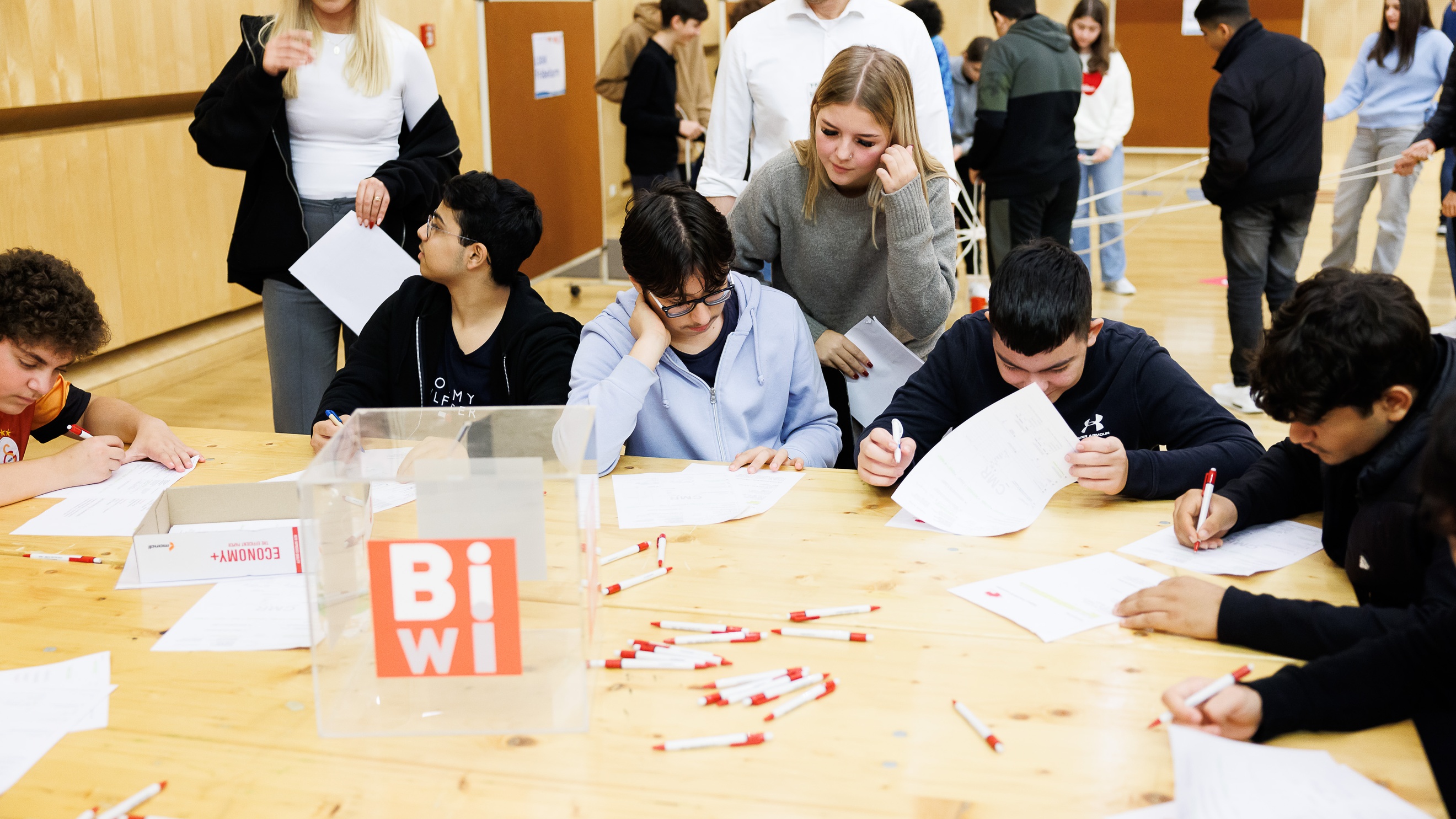 Um einen großen Tisch sitzt eine Gruppe von Teenagern. Jeder Teenager hat einen Zettel vor sich und schreibt darauf. Zwei Personen stehen neben dem Tisch