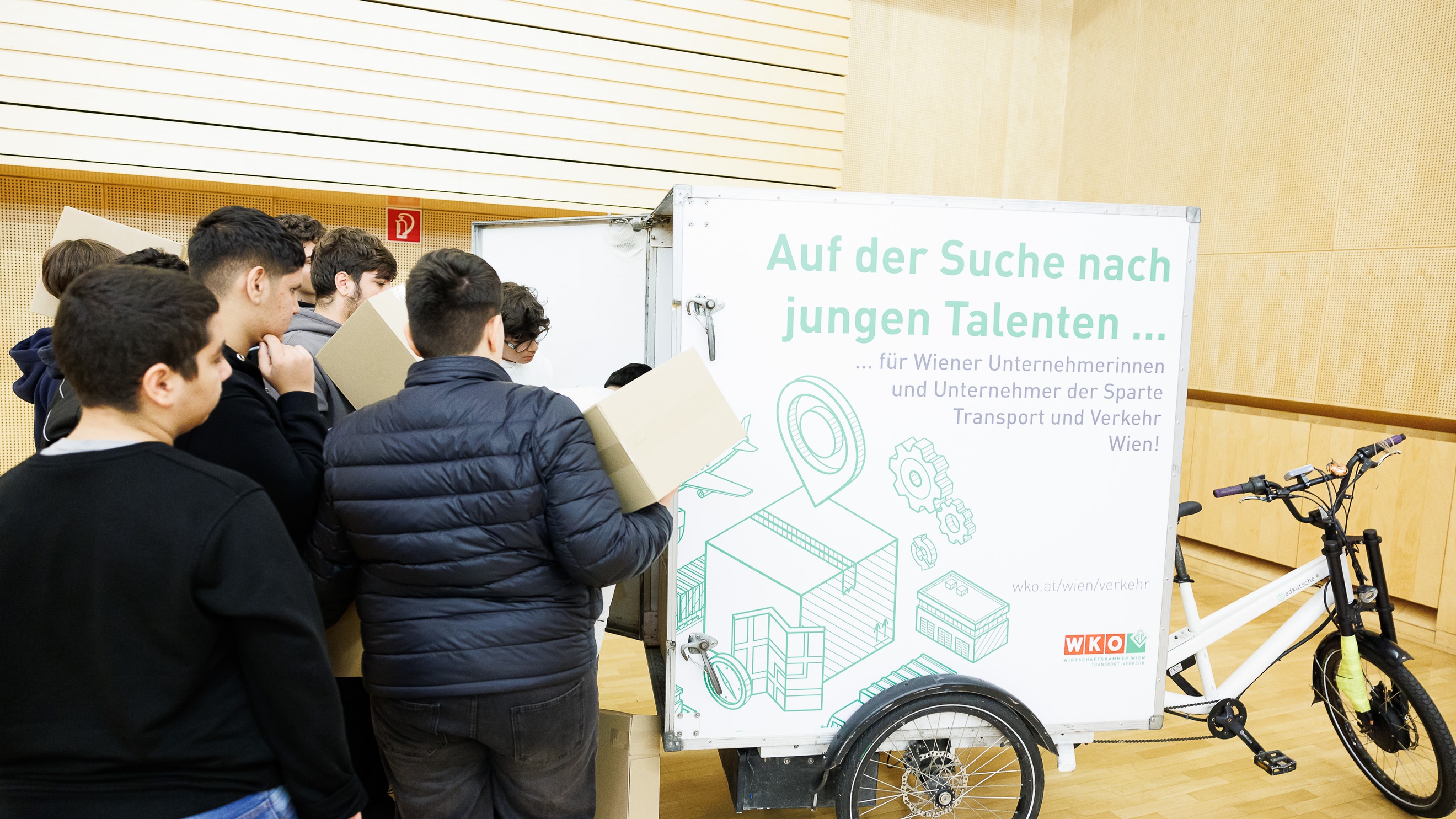 Hinter einem geschlossenen Anhänger, der an einem Fahrrad montiert ist, steht eine Gruppe von Personen. Ein paar von ihnen halten übereinander gestapelte Kartons
