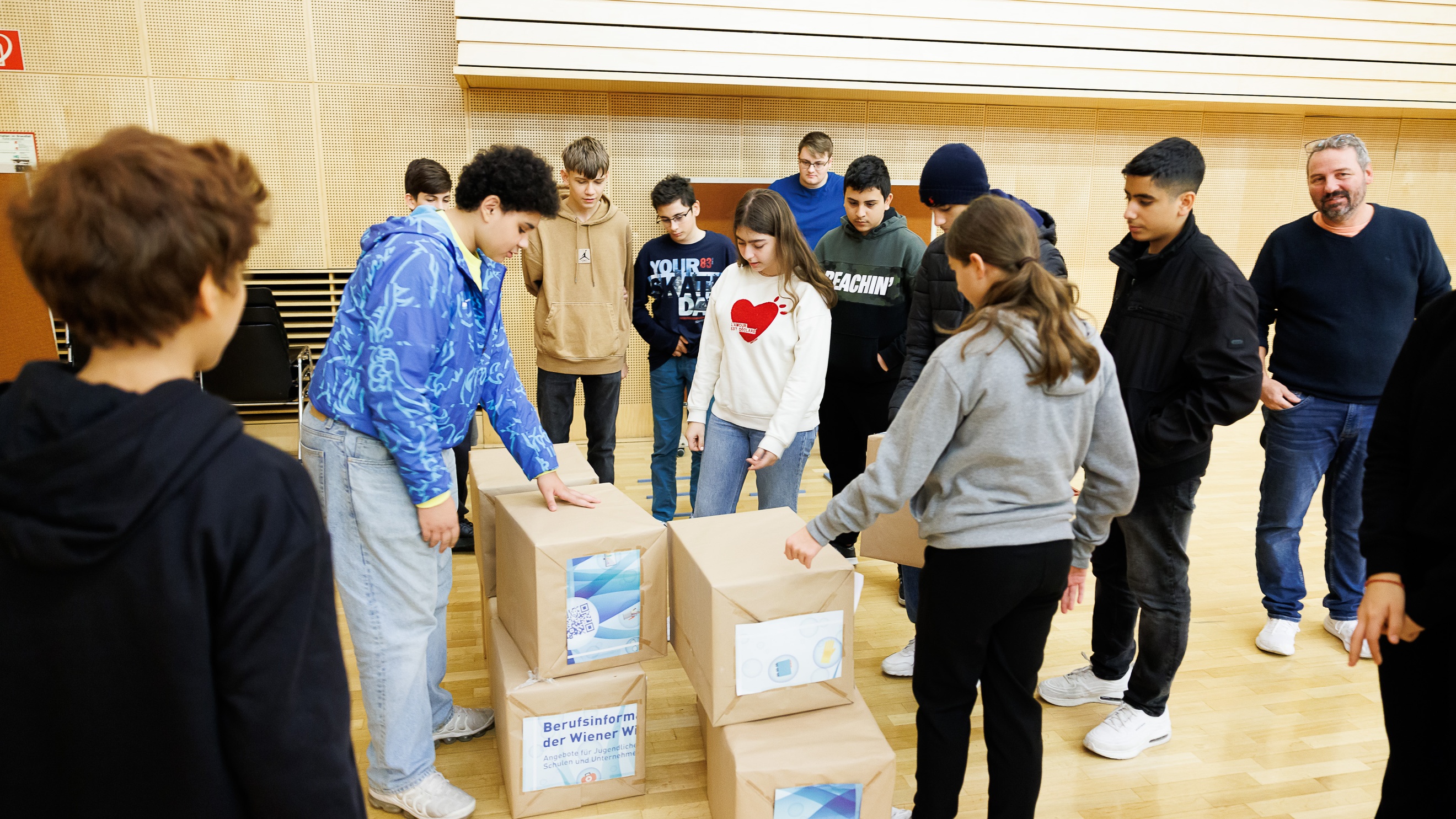 Am Boden stehen mehrere, in braunem Papier verpackte, rechteckige Objekte. Um diese steht eine Gruppe von Teenagern. Ein paar blicken auf die Objete