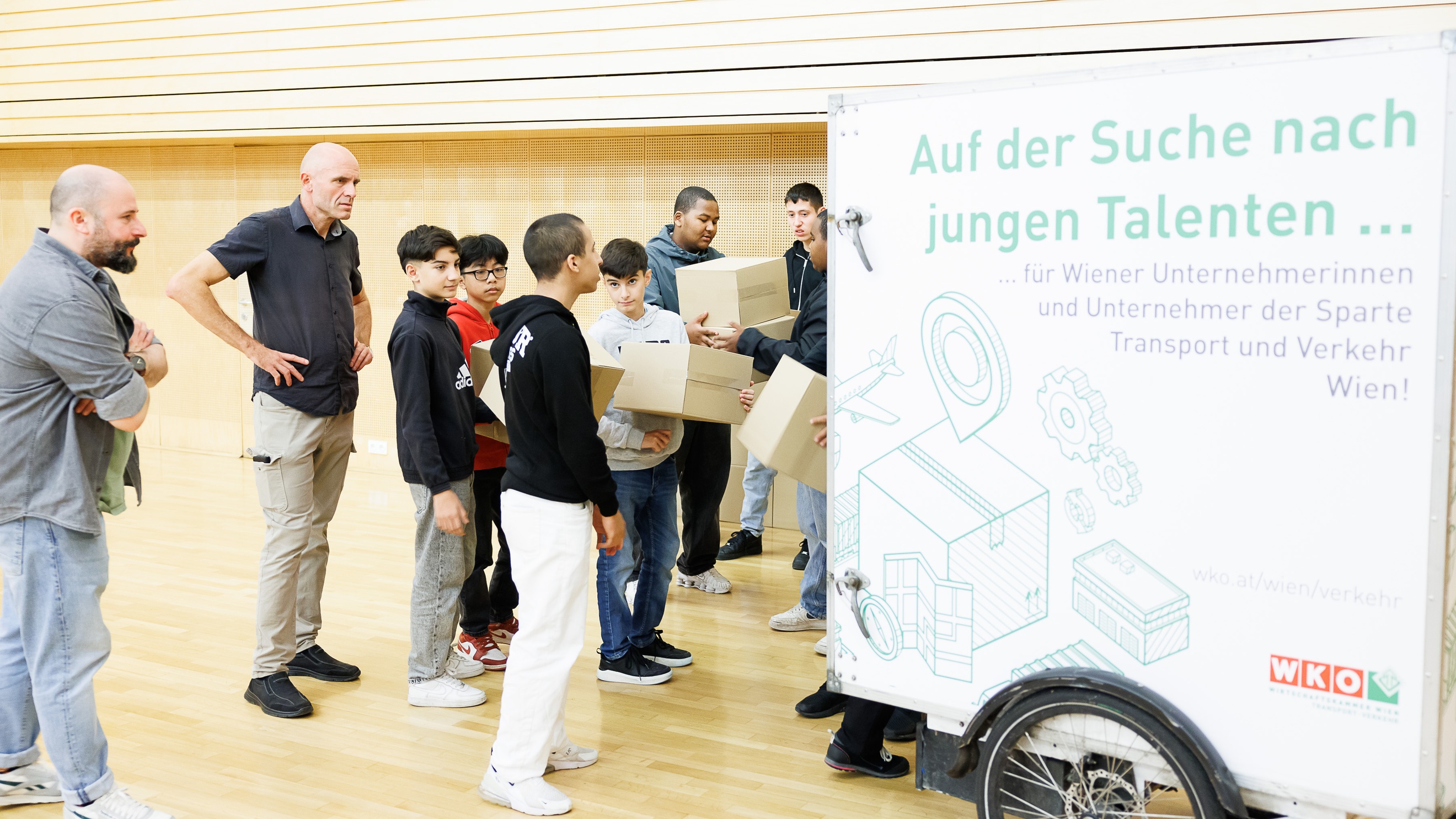 Hinter einem geschlossenen Anhänger, der an einem Fahrrad montiert ist, steht eine Gruppe von Personen. Ein paar von ihnen halten übereinander gestapelte Kartons