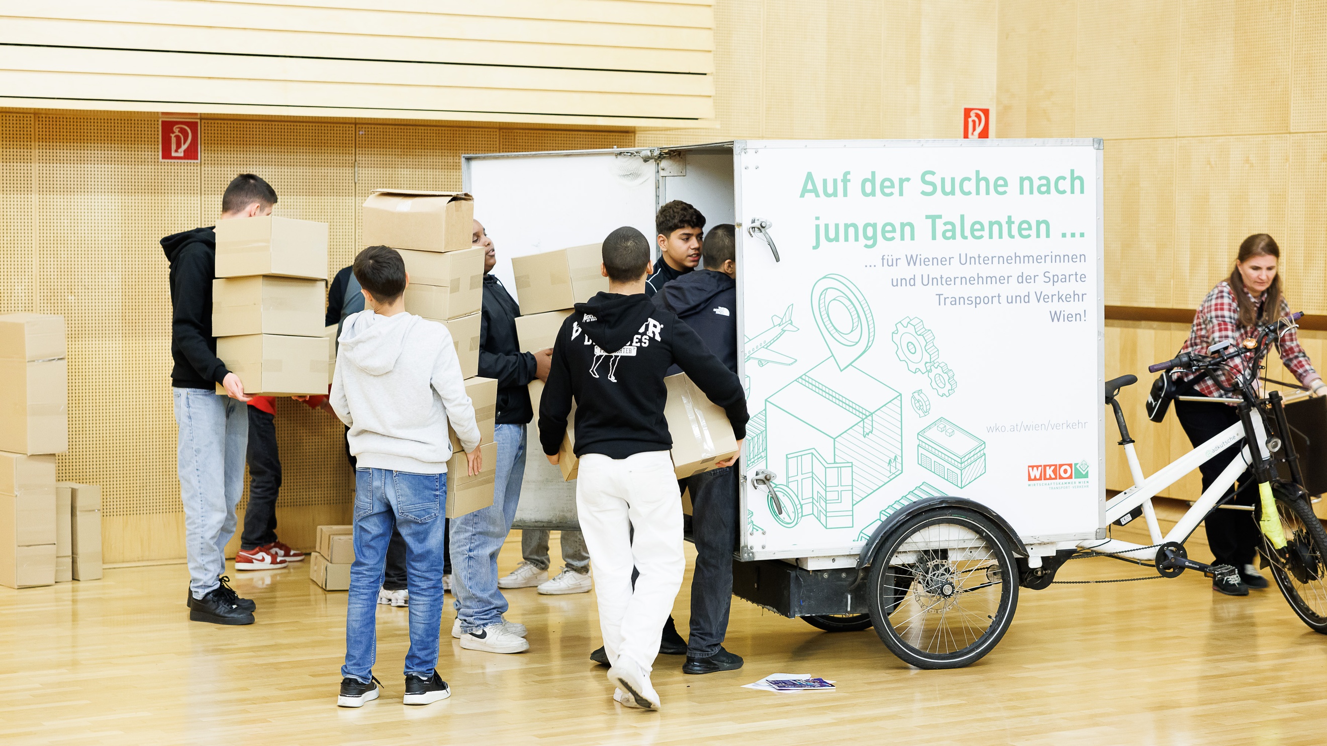 Hinter einem geschlossenen Anhänger, der an einem Fahrrad montiert ist, stehen sieben Personen, die alle einen Stapel Kartons halten