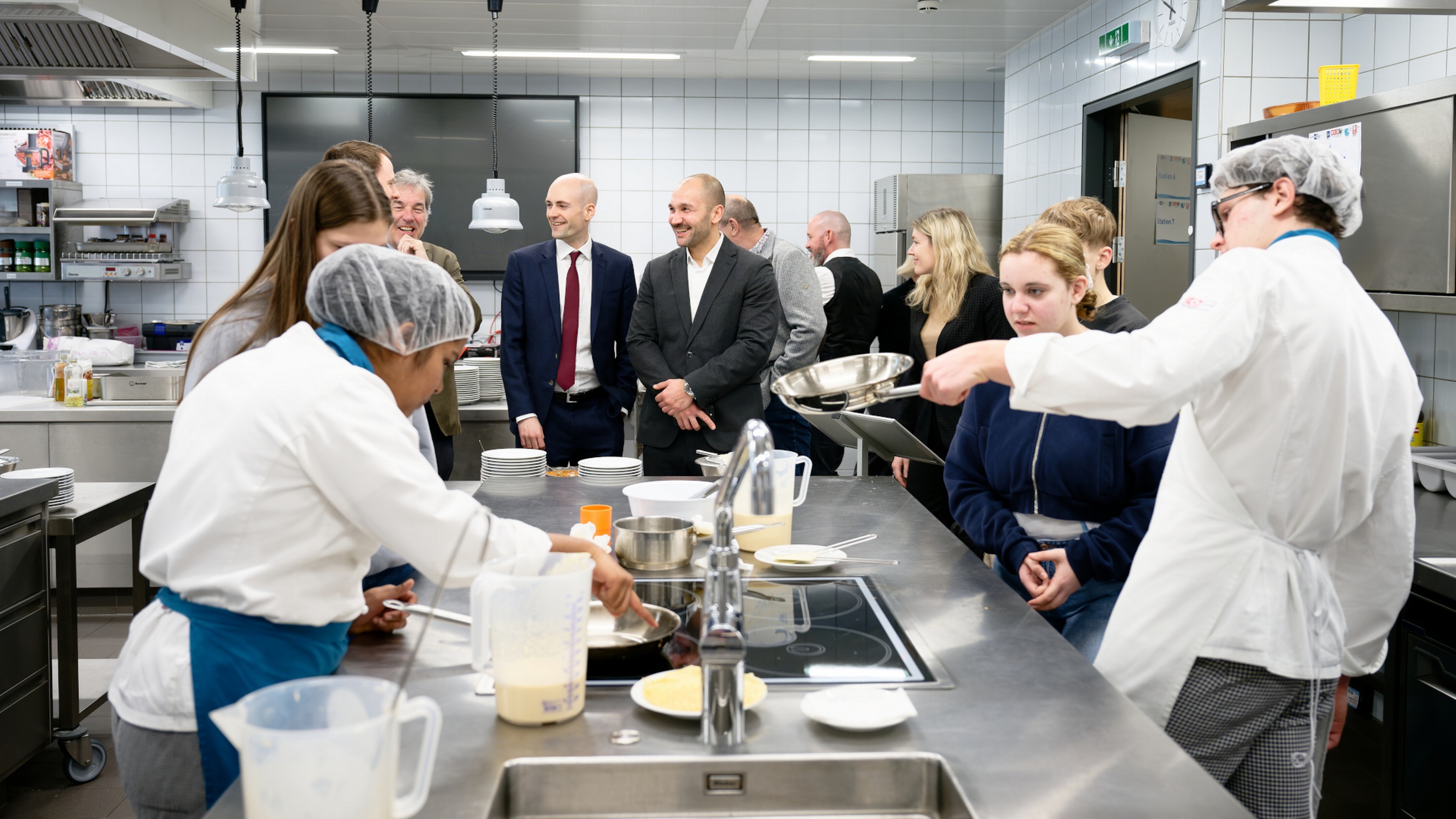 In einer Gastronomieküche stehen um einen Arbeitsblock zwei Personen mit Haarnetzen und Schürzen, die Jugendlichen, die neben ihnen stehen etwas erklären. Im Hintergrund sind weitere Jugendliche sowie Erwachsene, die sich unterhalten