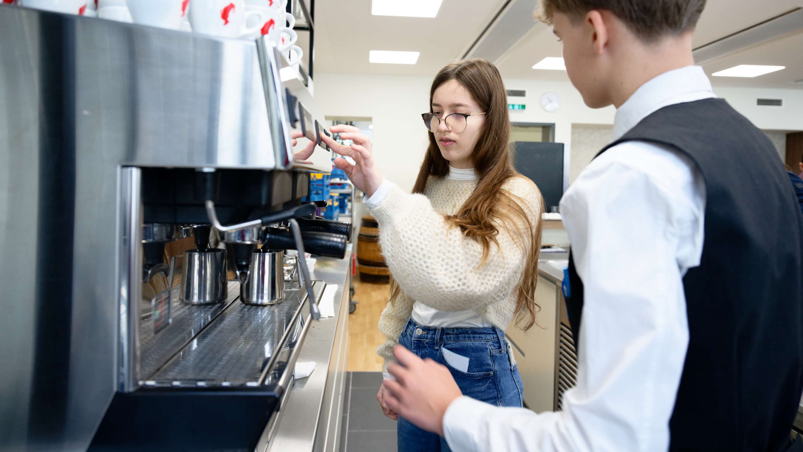 Neben einer Kaffeemaschine stehen eine jugendliche Person sowie ein Kellner. Die jugendliche Person drückt einen Knopf auf der Maschine